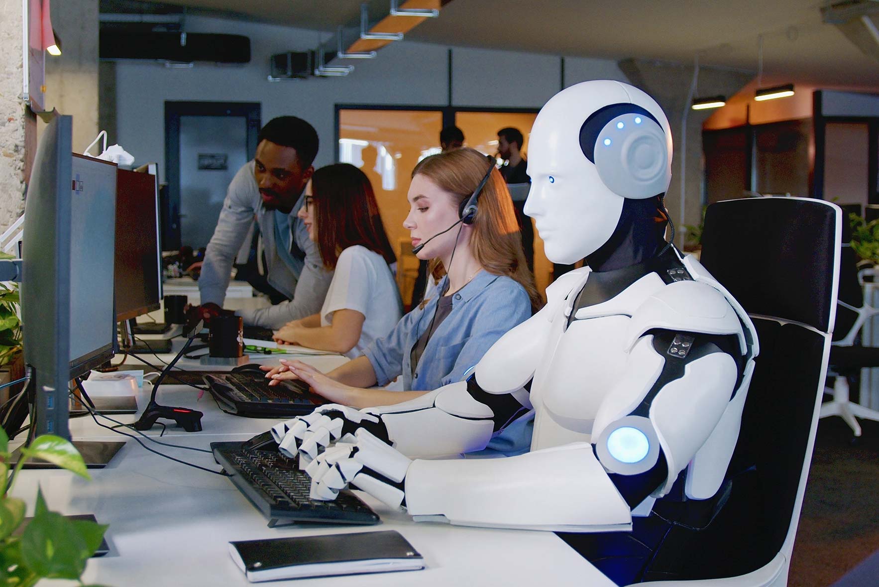 A futuristic office with a humanoid robot wearing a headset seated at a desk, typing on a keyboard next to a woman. Two colleagues are standing and discussing work in the background. Modern office environment with plants around.