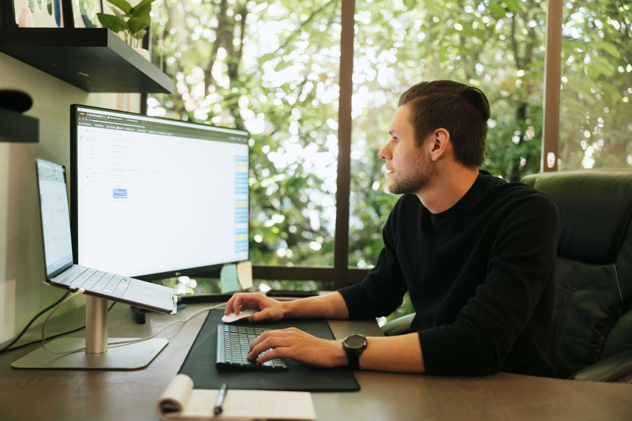 A person sits at a desk working on a computer with dual monitors. They wear a black shirt and are focused on the screen. The background shows a large window with greenery outside. A notebook and pen rest on the desk.