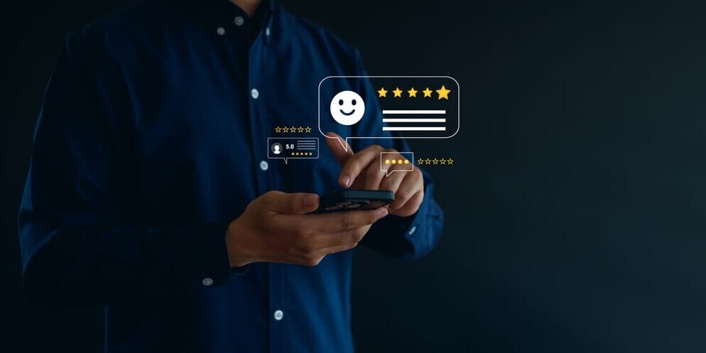 A person in a dark shirt uses a smartphone with holographic review icons above it, including a smiling face and five stars, suggesting satisfaction or positive feedback. Dark background enhances the glowing digital elements.