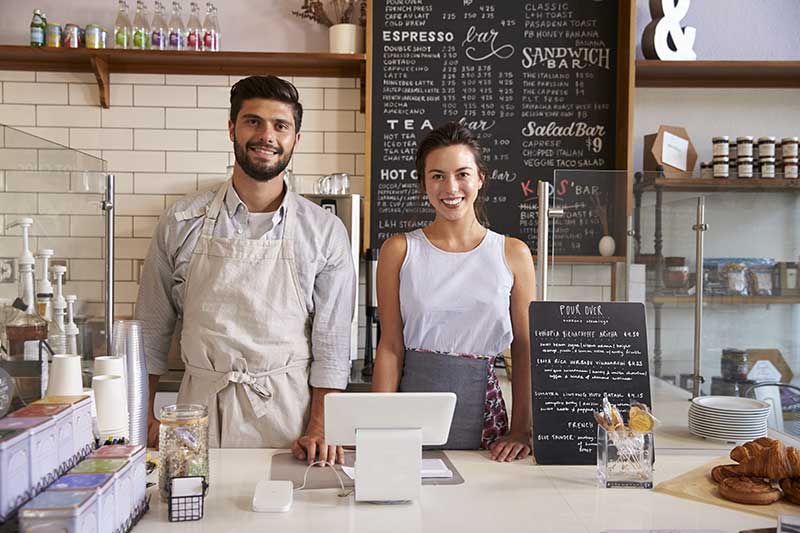 Two people standing behind a counter in a cafe. They are smiling and wearing aprons. Behind them is a chalkboard menu with various drink and food options. Cookies and other items are displayed on the counter.