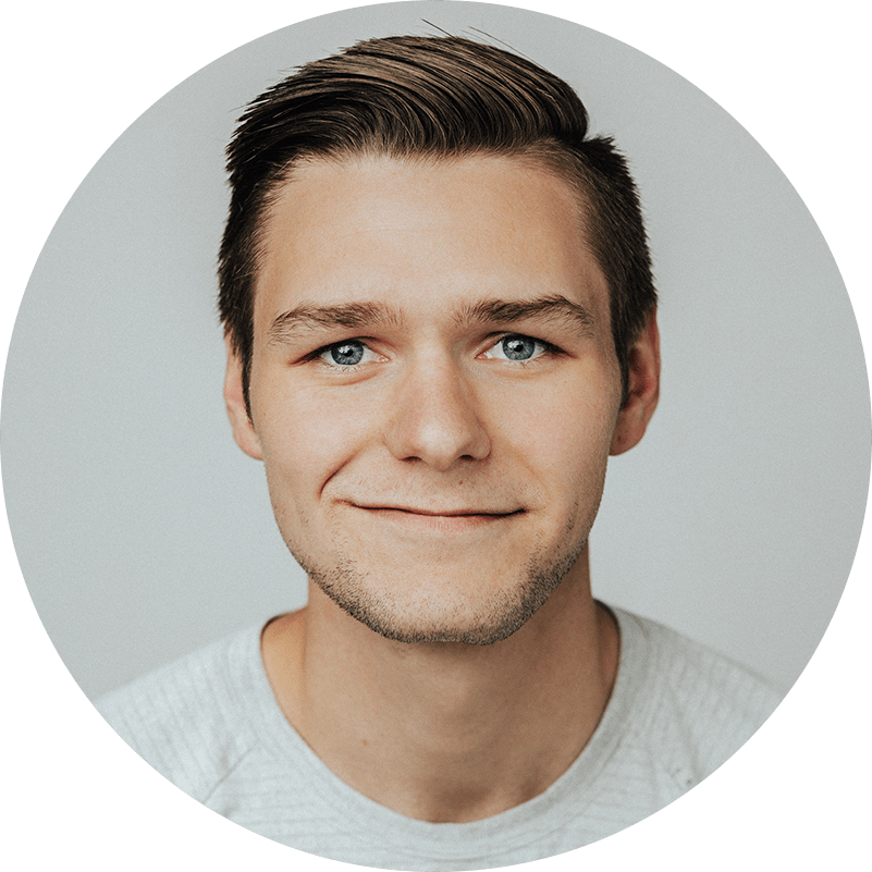 A person with short brown hair and blue eyes smiles slightly, wearing a light gray shirt. The background is plain and light-colored.