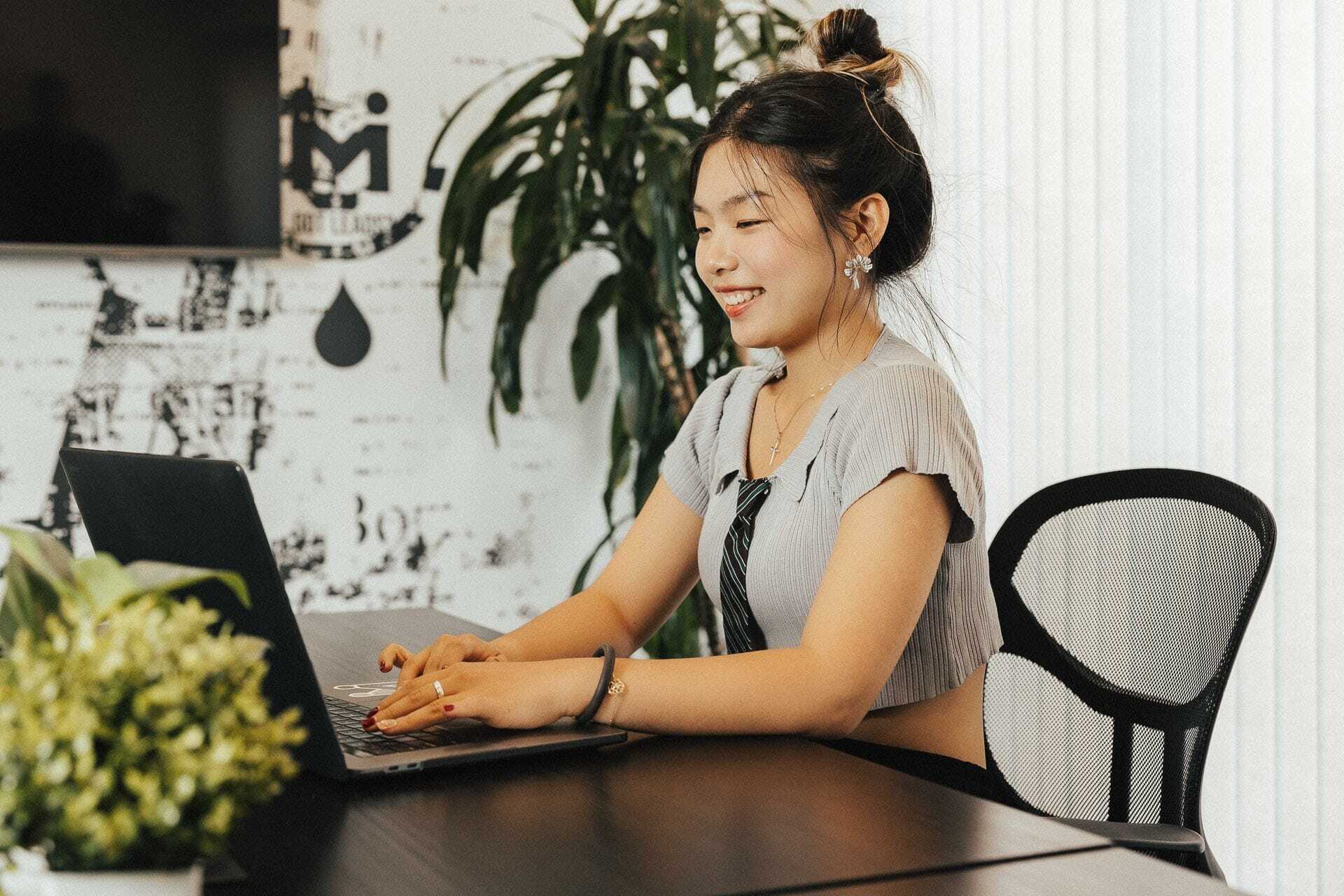 A woman is sitting at a desk, smiling as she types on a laptop. She is wearing a short-sleeved top and tie. A potted plant and a logo on the wall are in the background. Sunlight streams through vertical blinds.