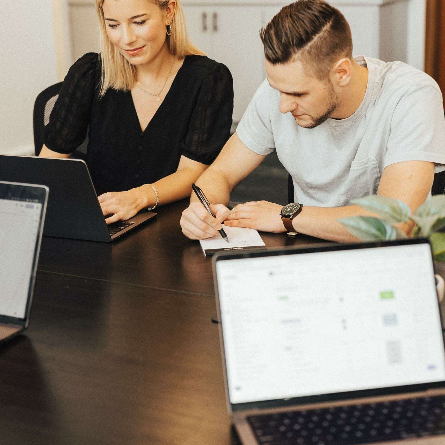 Two people working at a table with laptops. A woman is typing on her laptop, while a man takes notes on paper. Other laptops are visible on the table, along with a small plant in the foreground.