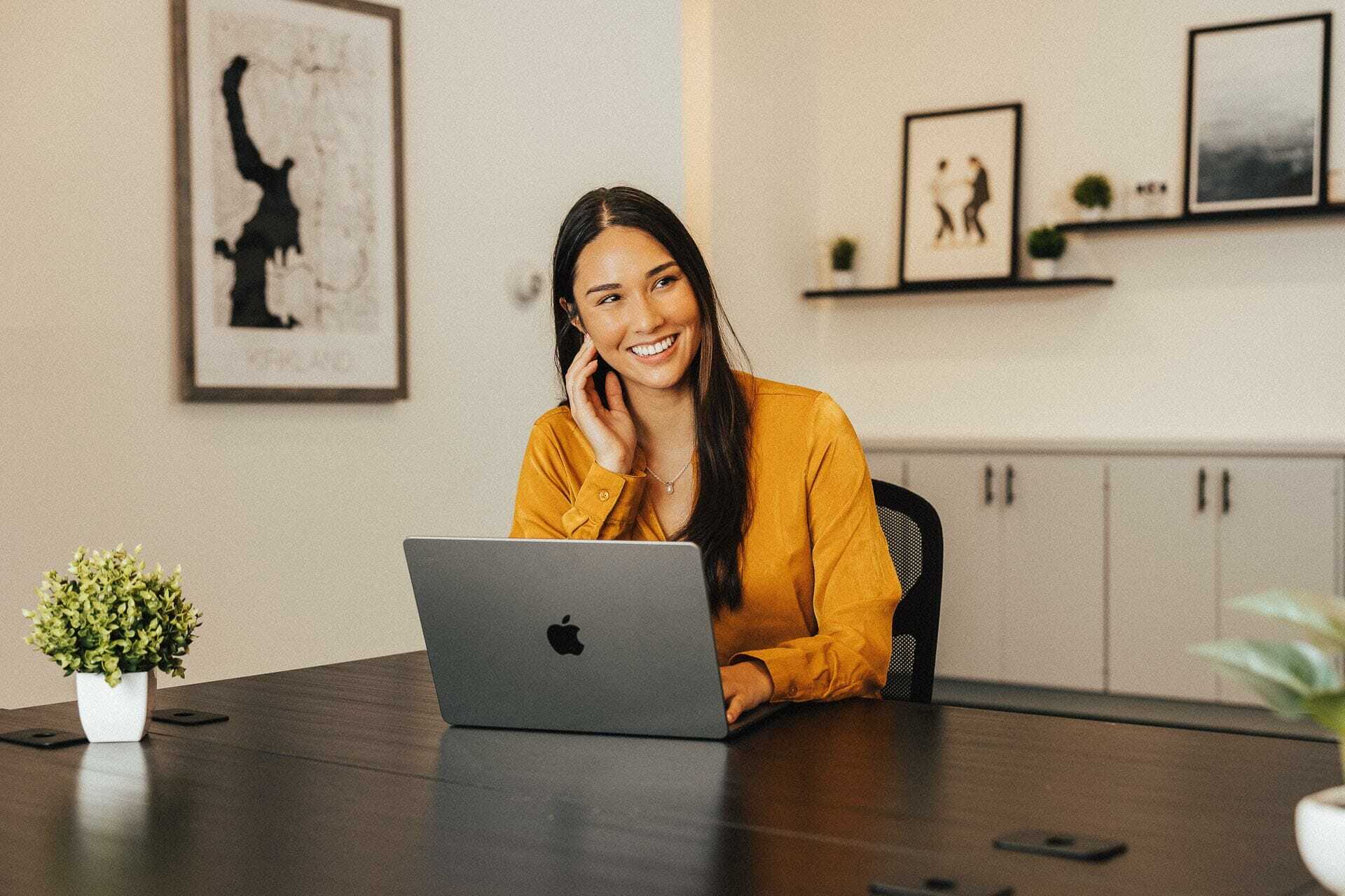 A woman with long dark hair in a yellow shirt smiles while sitting at a table with a laptop. The room has framed art on the walls and small potted plants on the table.