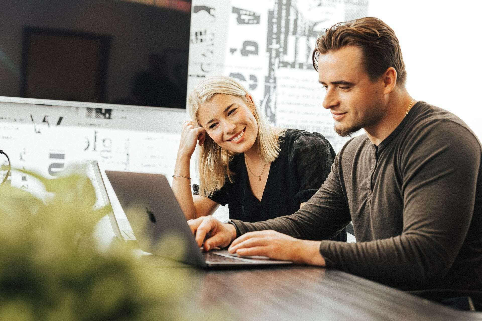 Two people sitting at a table, using laptops. The woman on the left is smiling and looking at the man on the right, who is focused on his screen. The room has a bright and modern decor with some wall art in the background.