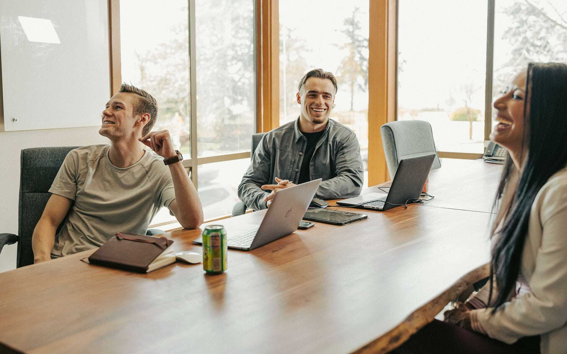 Three people sitting at a conference table with laptops, smiling and laughing. A window in the background shows a bright, sunny day. A can of drink and a notebook are on the table.