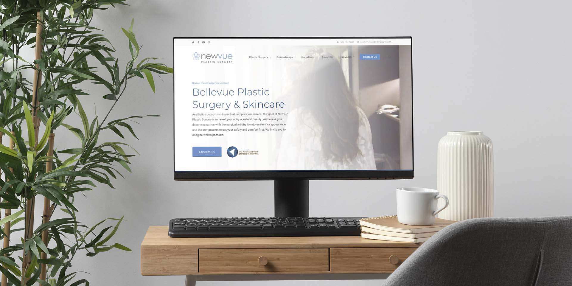 A computer monitor on a wooden desk displays a website for Bellevue Plastic Surgery & Skincare. On the desk are a potted plant, a stack of books, and a white vase. A gray chair is partially visible.