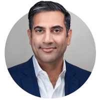 Portrait of a person with short dark hair, wearing a navy suit and white shirt, smiling slightly against a gray background.