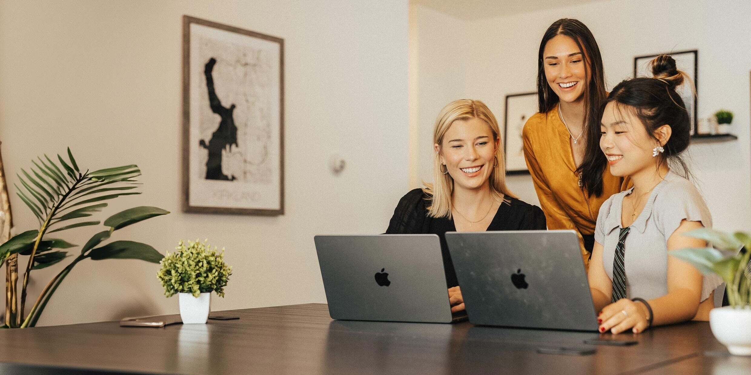 Three women are sitting at a table working on laptops, smiling and engaged in a discussion. The room is decorated with plants and framed artwork. The atmosphere is bright and collaborative.