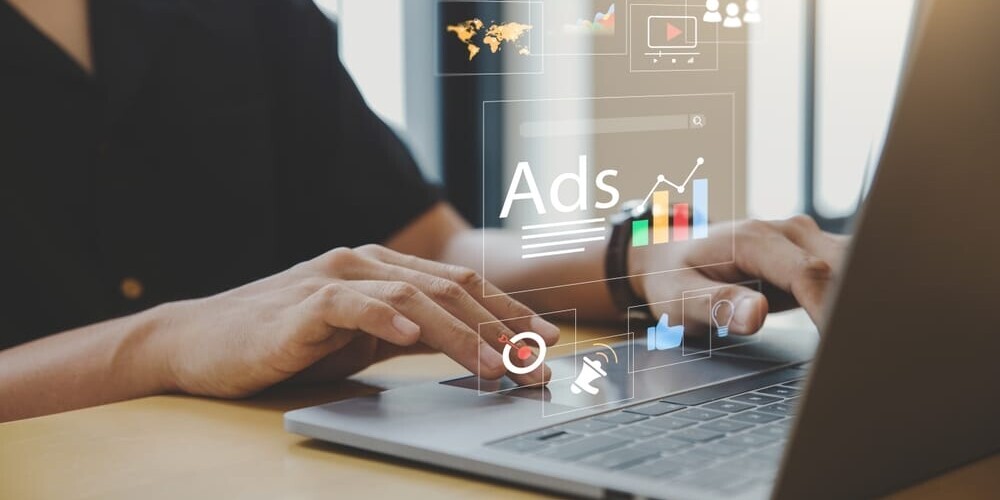 Person typing on a laptop with digital marketing icons floating above the keyboard, including graphs, a globe, a bell, a thumbs-up, and the word Ads. The scene is well-lit, suggesting a focus on online advertising and analytics.