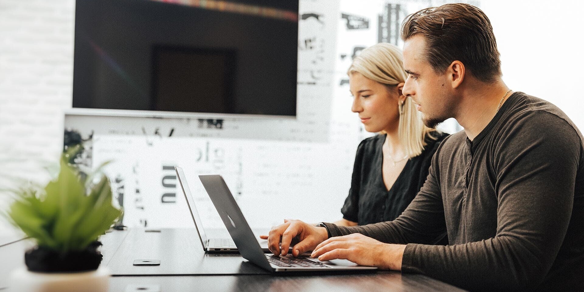 A man and woman are sitting at a table using laptops. They appear to be working or collaborating in a modern office setting, with a large TV screen in the background and a potted plant on the table.