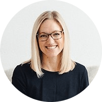 A person with long, straight blonde hair and glasses smiles while looking at the camera. They are wearing a black top and are seated against a plain white background.