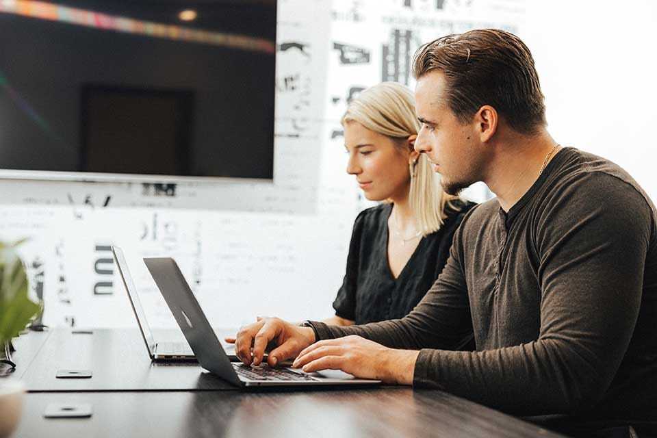 Two people sit at a table working on laptops. A man types on his laptop while a woman looks at her screen. They are in a bright room with a large screen on the wall in the background.