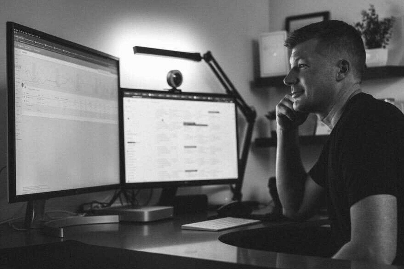 A person in a black t-shirt is sitting and smiling at a desk with two large computer monitors displaying charts and data. The room is dimly lit, with a sleek, modern design, and features a plant and shelves in the background.