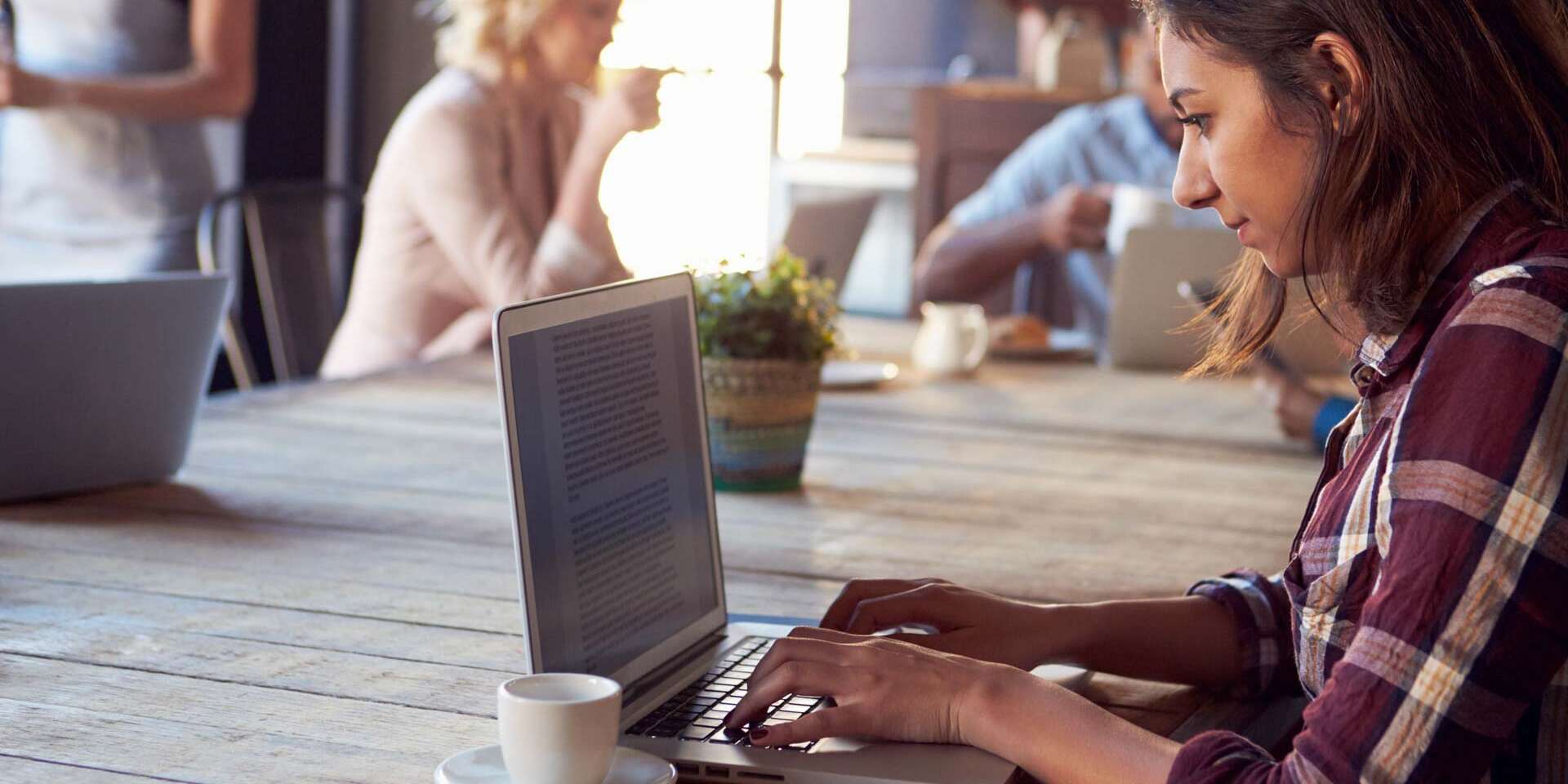 A woman in a plaid shirt types on a laptop at a wooden table in a café. A cup of coffee sits beside her. In the background, two people are conversing, and another person is drinking from a mug. A small plant decorates the table.