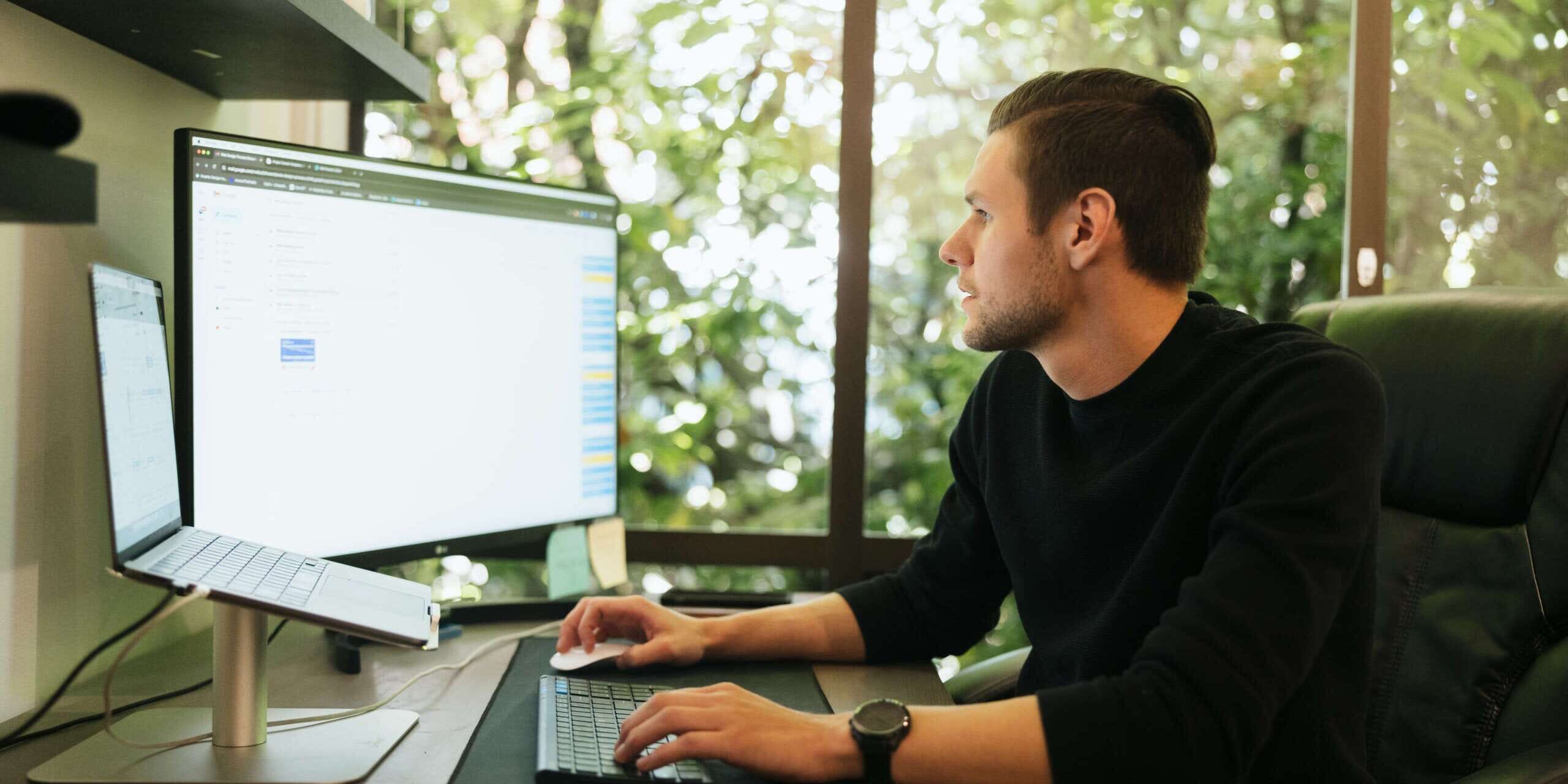 A person sits at a desk working on a computer with dual monitors. They wear a black shirt and are focused on the screen. The background shows a large window with greenery outside. A notebook and pen rest on the desk.