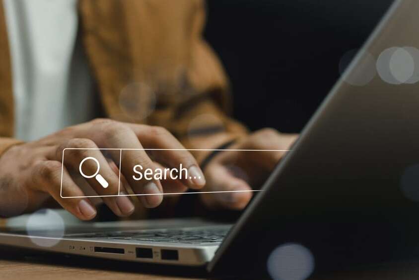Person typing on a laptop with a transparent search bar overlay, featuring a magnifying glass icon next to the word Search.... The background is blurred, focusing on the hands and keyboard.