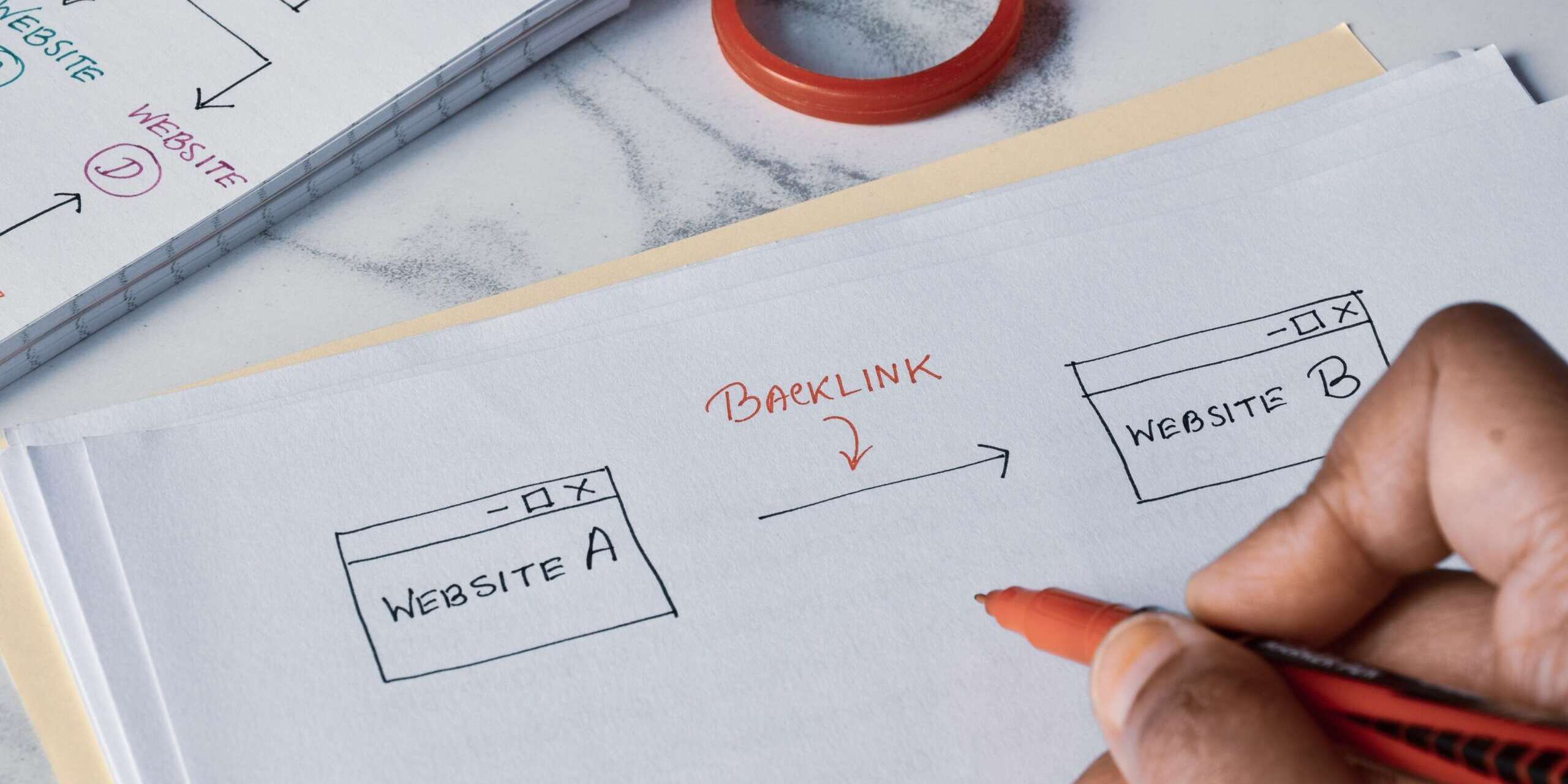 A person sketches a diagram on paper using a red pen, illustrating a backlink from Website A to Website B. Nearby, a magnifying glass and a notebook filled with similar drawings rest on a marble surface.