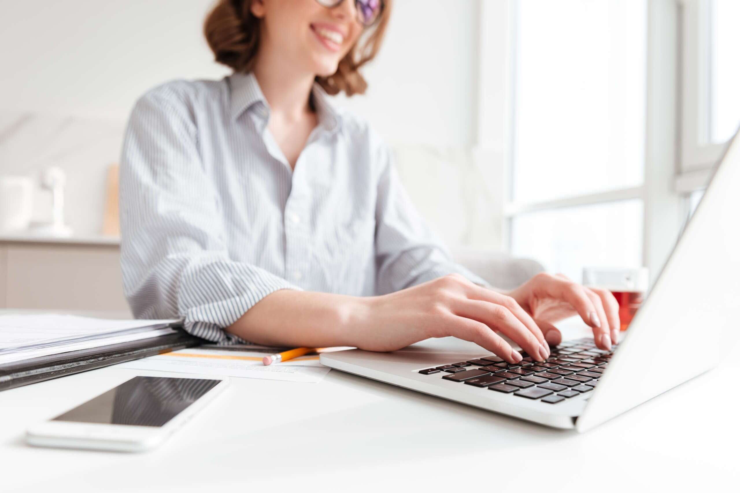 A woman in a striped shirt is smiling while typing on a laptop at a white desk. The desk also has a smartphone, a pen, and a notebook. Sunlight streams through a window in the background.