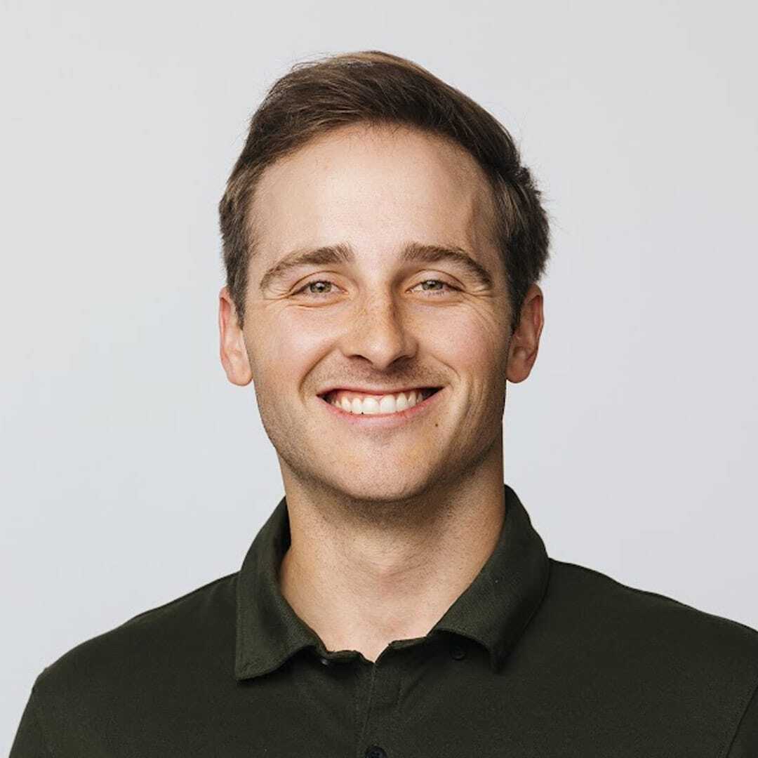 A young man with short brown hair smiles at the camera. He is wearing a dark green collared shirt and standing against a plain, light-colored background.