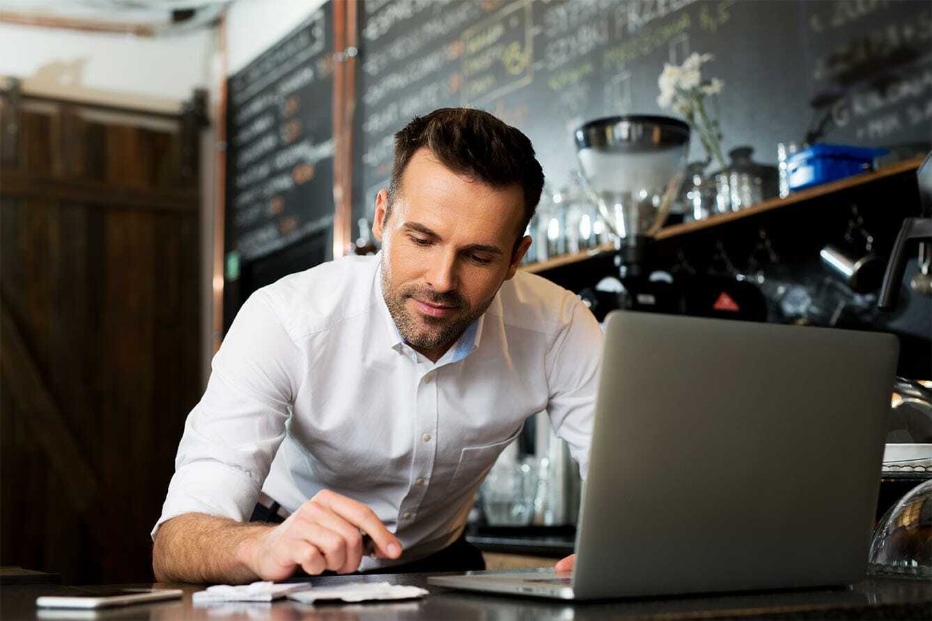 Businessman in a white shirt leans over a table, using a laptop and checking documents. A chalkboard with writing is in the background, suggesting a cozy cafe or office setting.