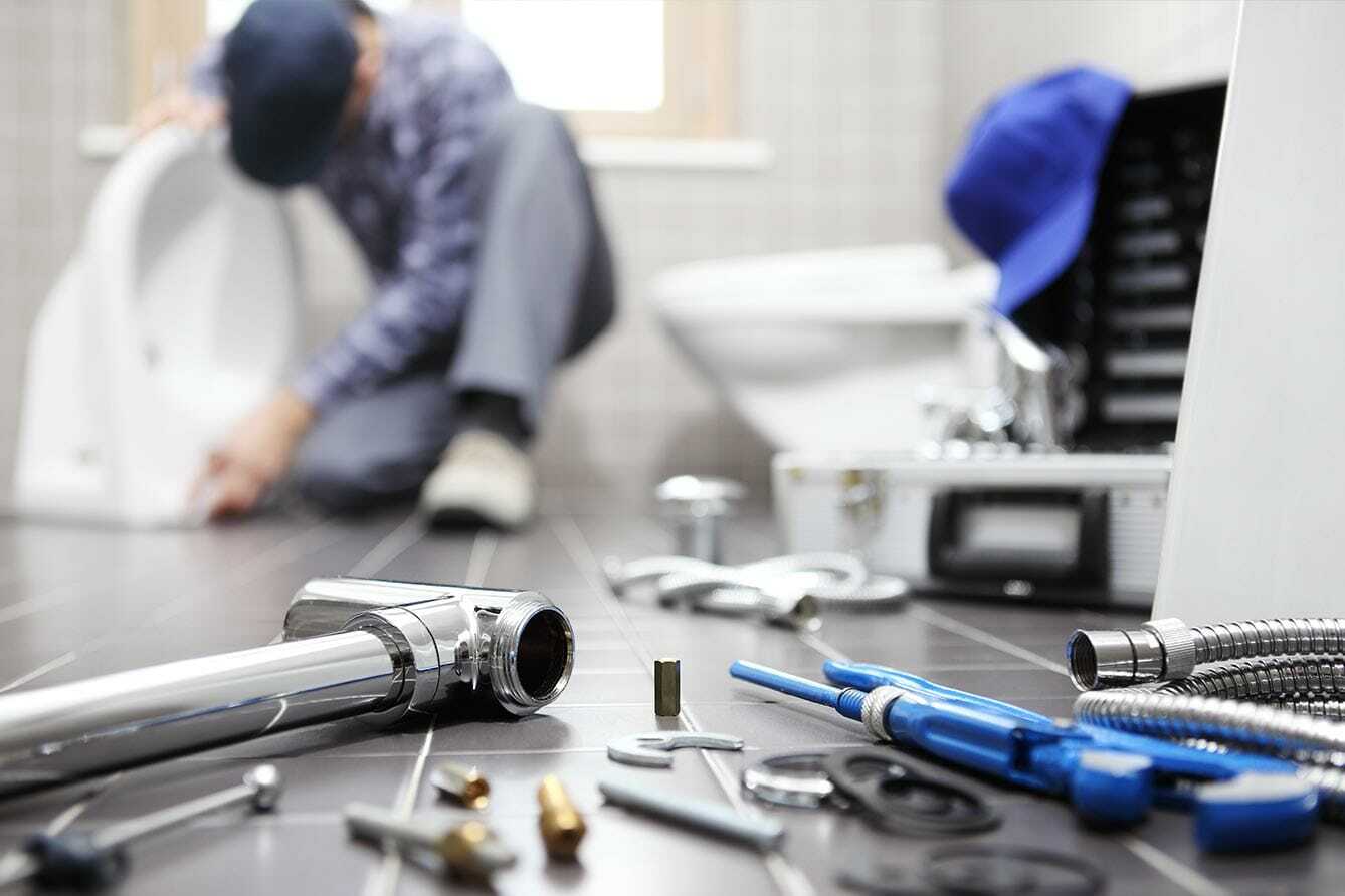 A plumber is kneeling and working on a toilet in a bathroom. Various tools and plumbing parts are scattered on the tiled floor, with a toolbox open nearby. A blue cap rests on a toilet seat in the background.