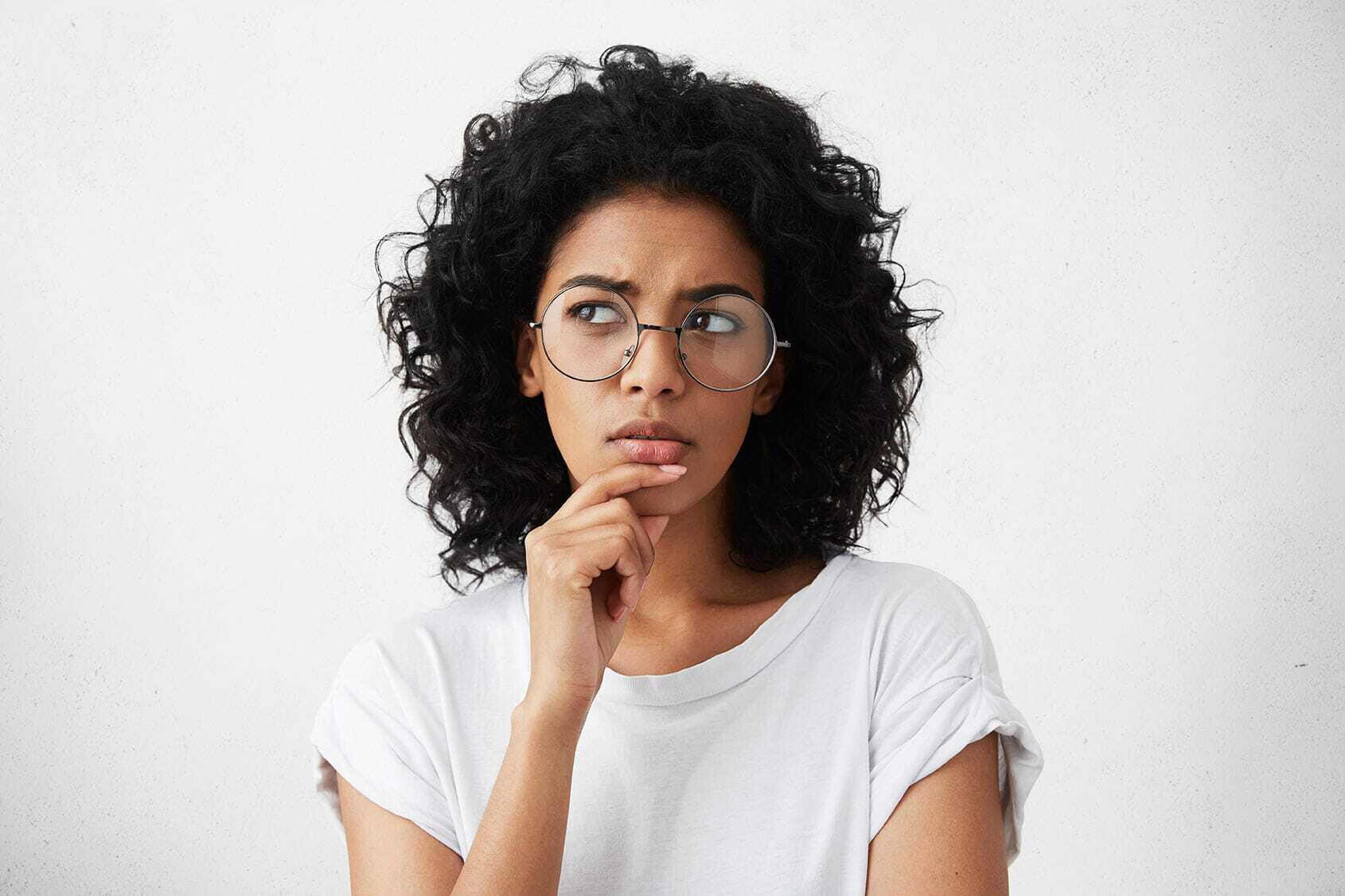 Person with curly hair and glasses wearing a white shirt, looking thoughtful with a slight frown. Their hand is placed on their chin, and they are gazing slightly to the side against a plain background.