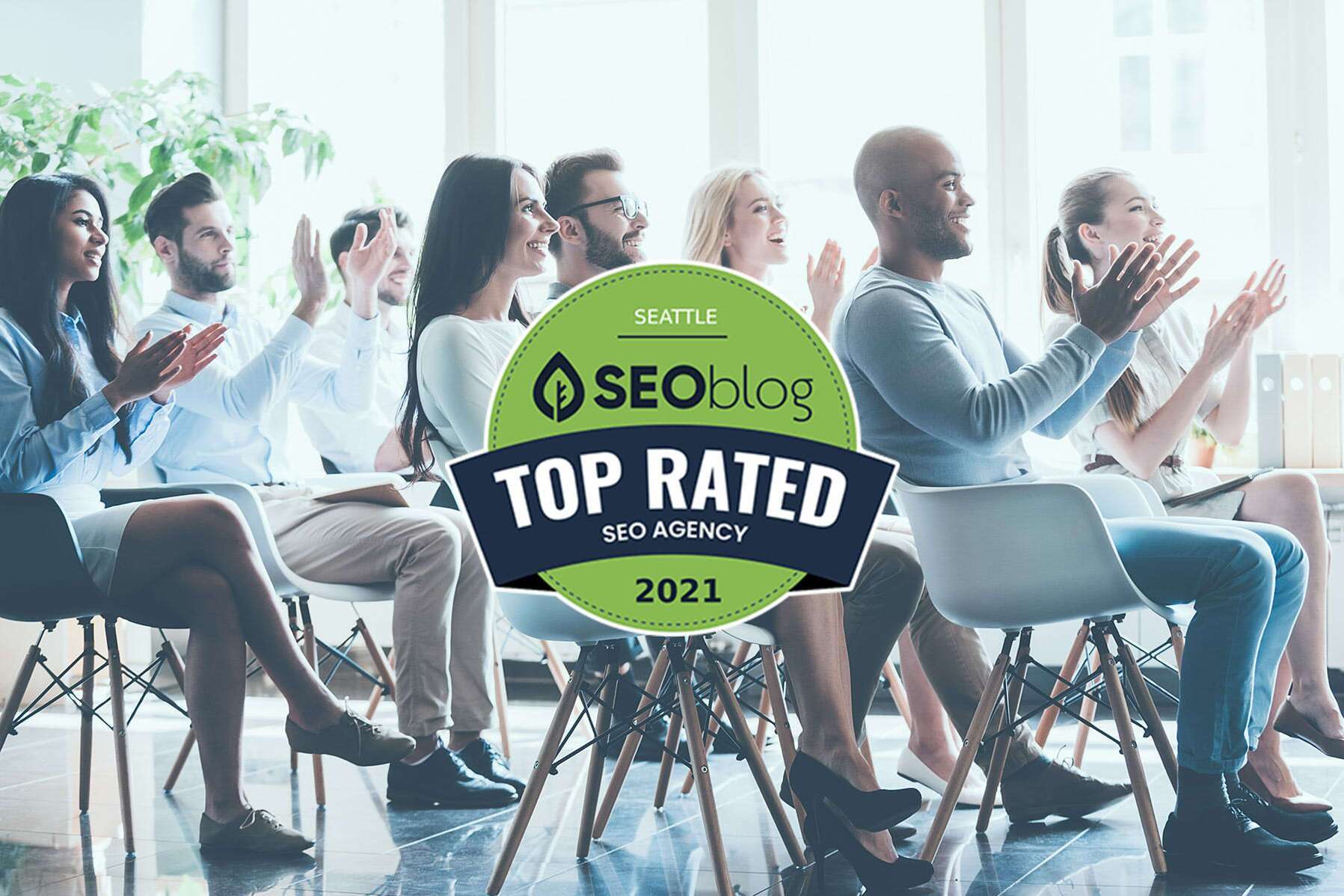 A diverse group of people sitting in chairs applauding. In the foreground is a badge reading Seattle SEOblog Top Rated SEO Agency 2021. The room is bright with large windows and plants in the background.