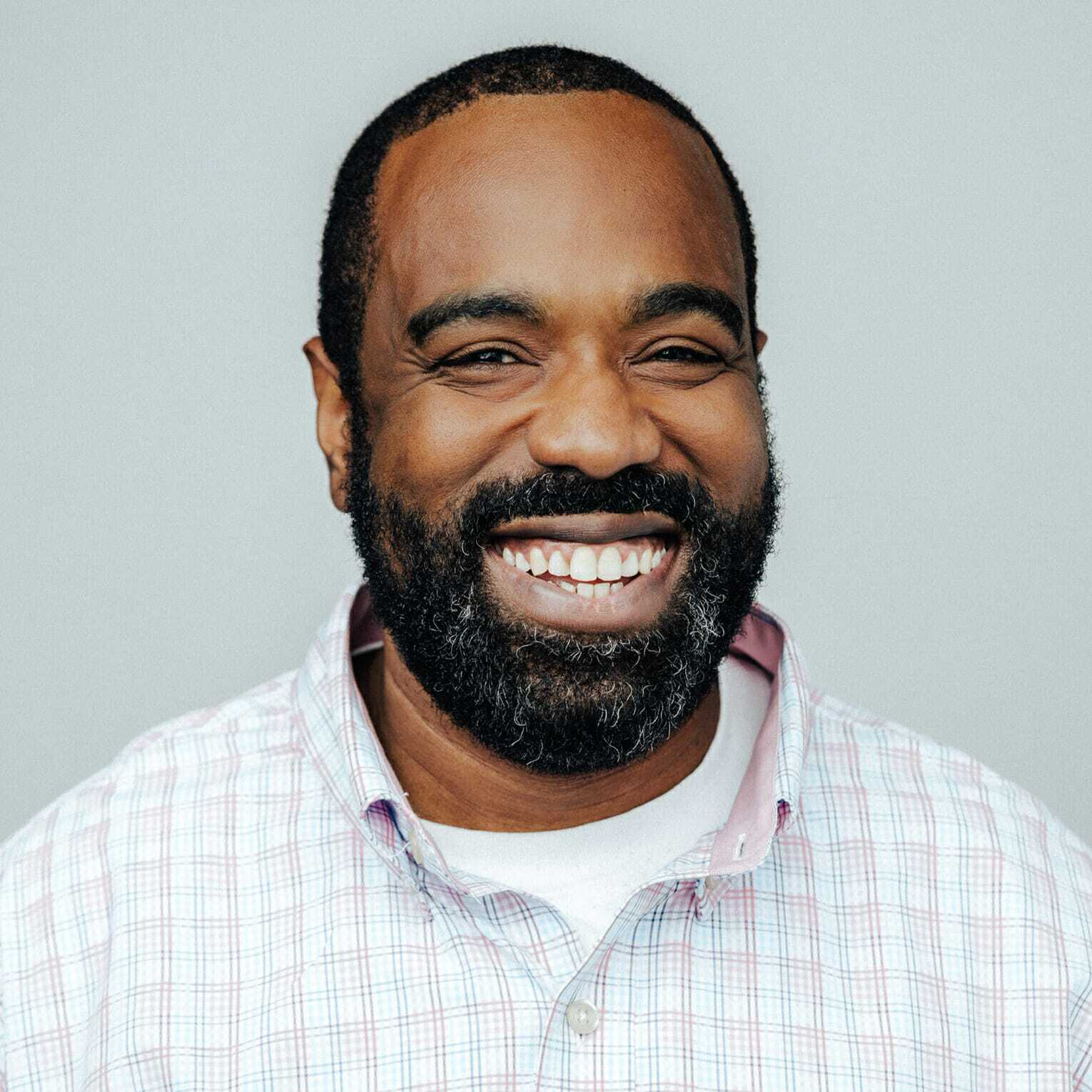 A man with a beard is smiling broadly. He is wearing a white and pink checkered shirt with a white undershirt against a plain gray background.