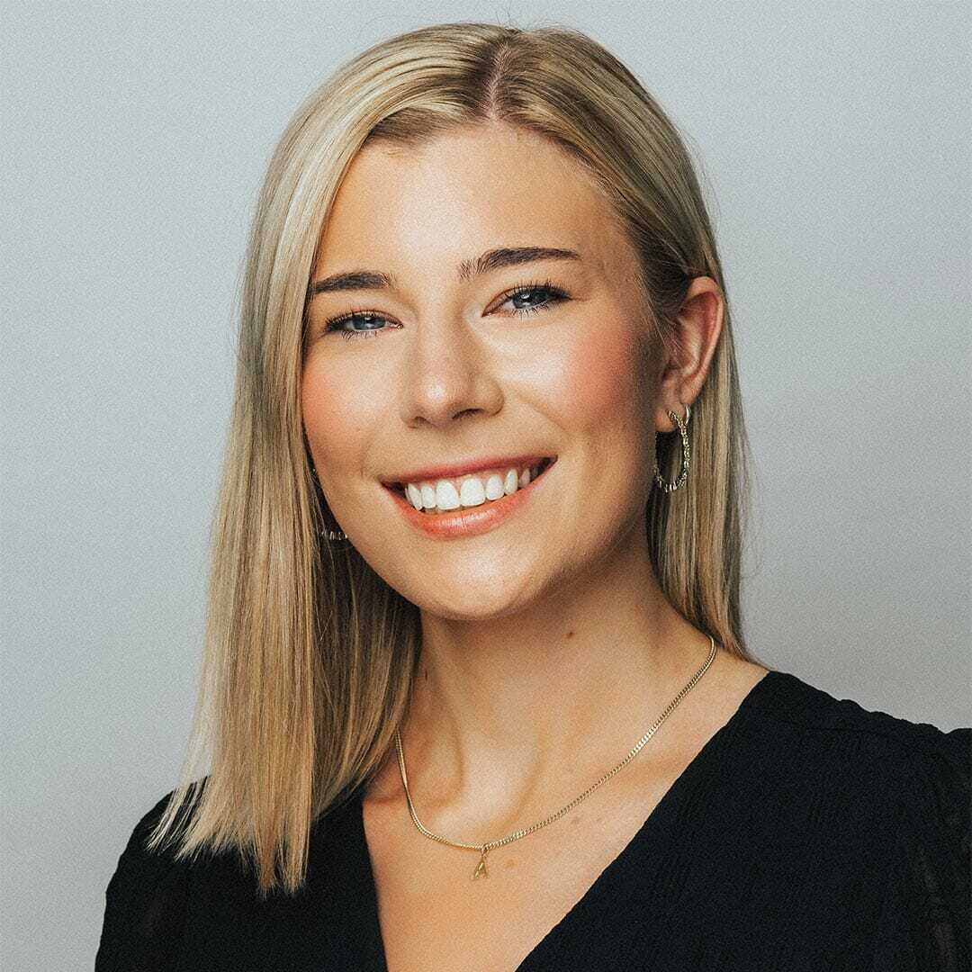 Smiling woman with straight blonde hair wearing a black top, hoop earrings, and a necklace, against a plain background.