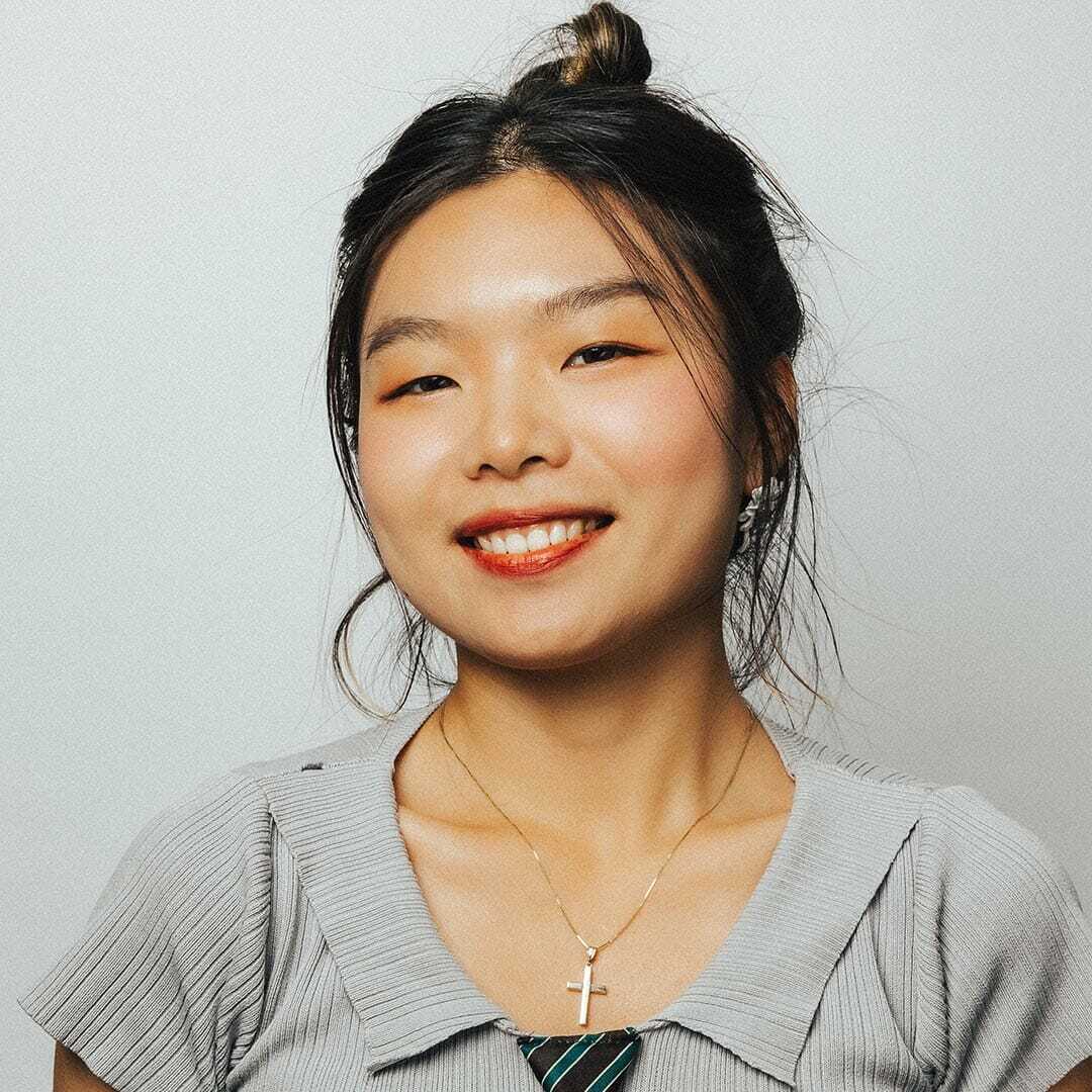 A person with long hair tied up, wearing a light gray top and a necklace with a cross pendant, smiles against a plain background.