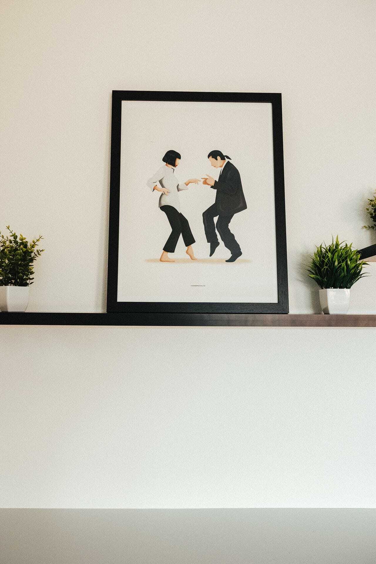 A framed poster on a shelf depicts two people dancing. The person on the left wears a white shirt and hat, and the person on the right is in a black suit. Two small potted plants are on either side of the frame.