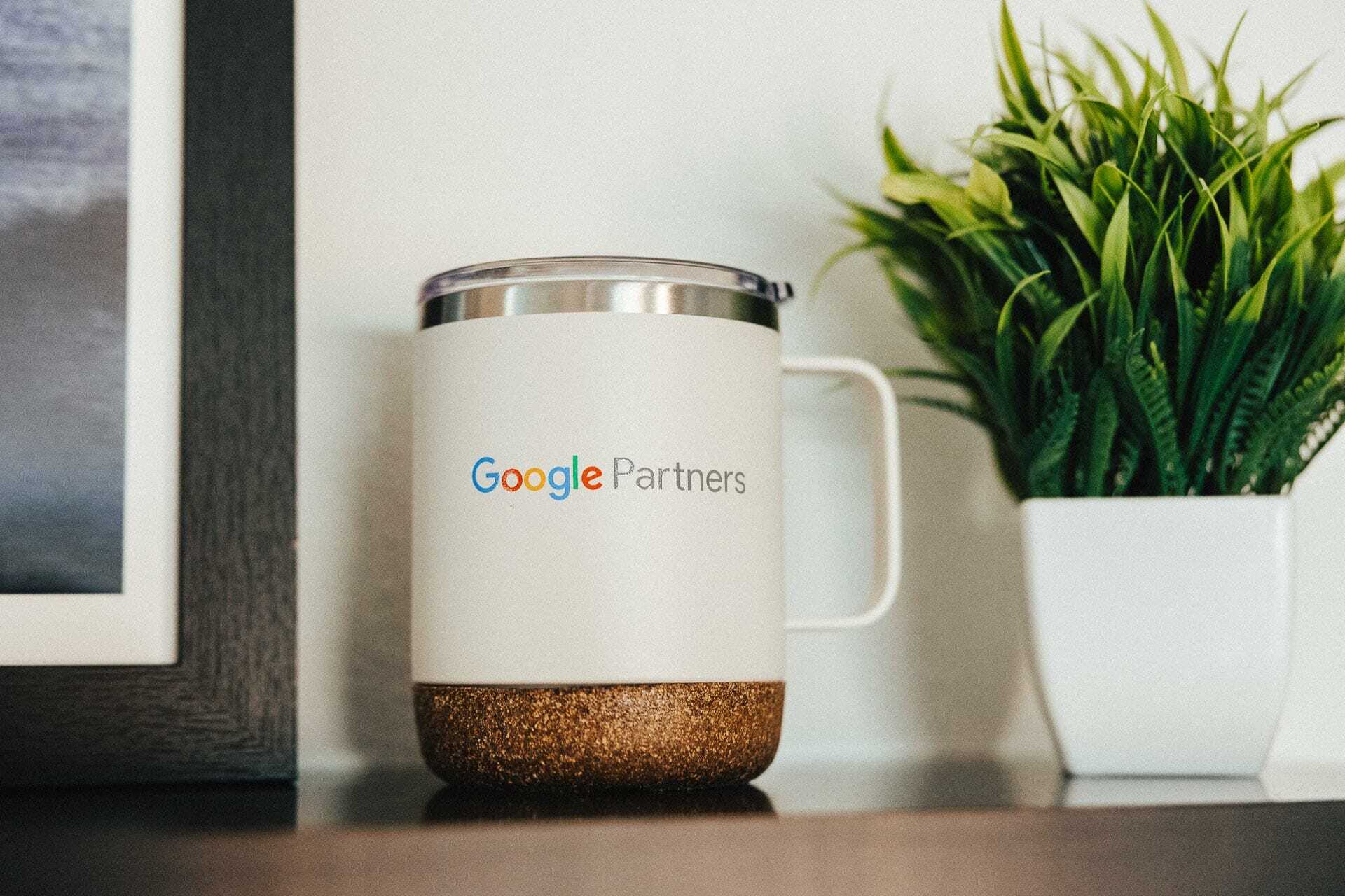 A white mug with Google Partners printed on it, featuring a metallic lid and a textured base, is placed next to a small potted plant with green leaves on a shelf.