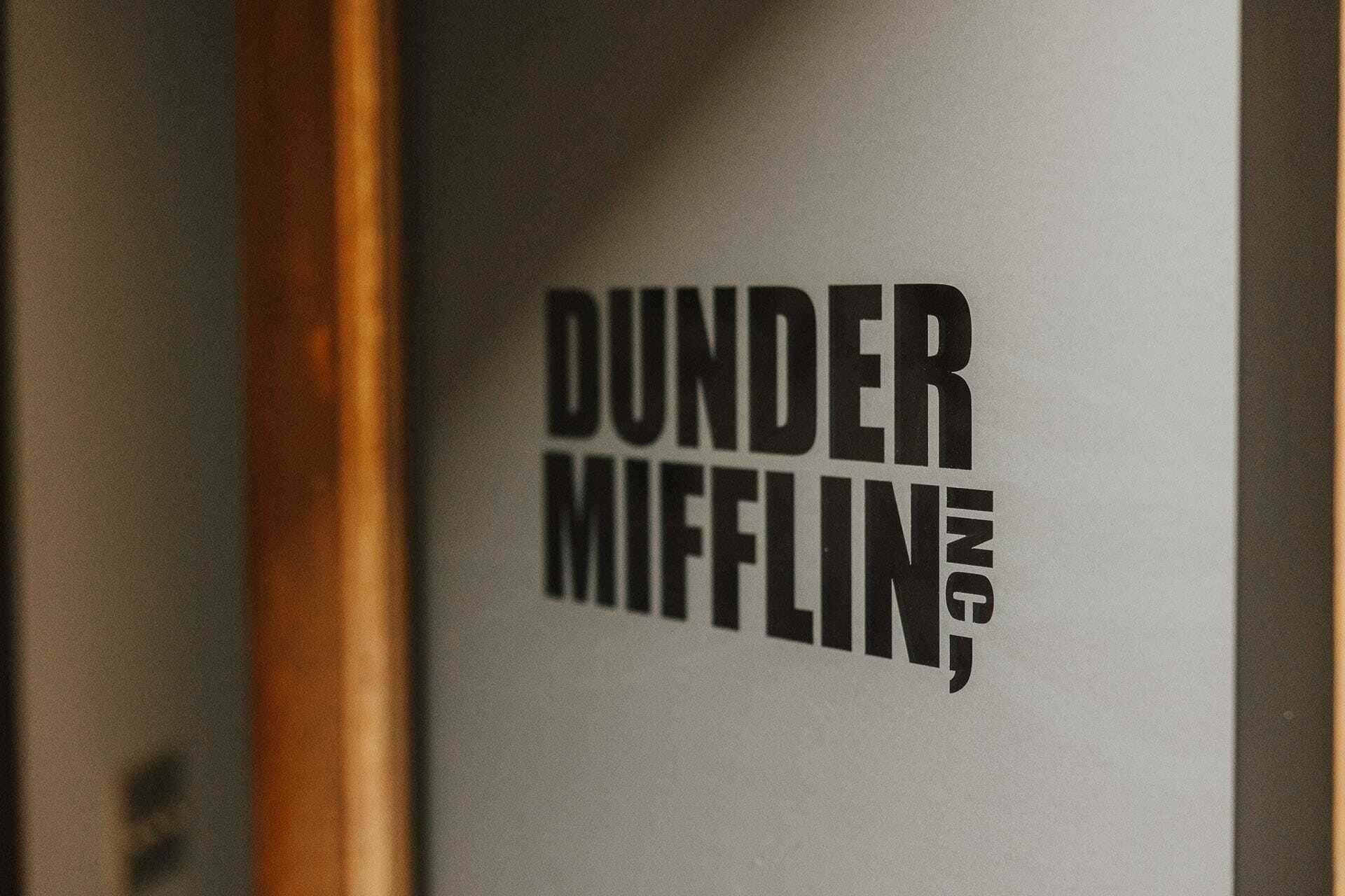 Close-up view of a grey wall with bold black text that reads Dunder Mifflin, Inc. The lighting creates a subtle shadow behind the letters, suggesting depth and texture.