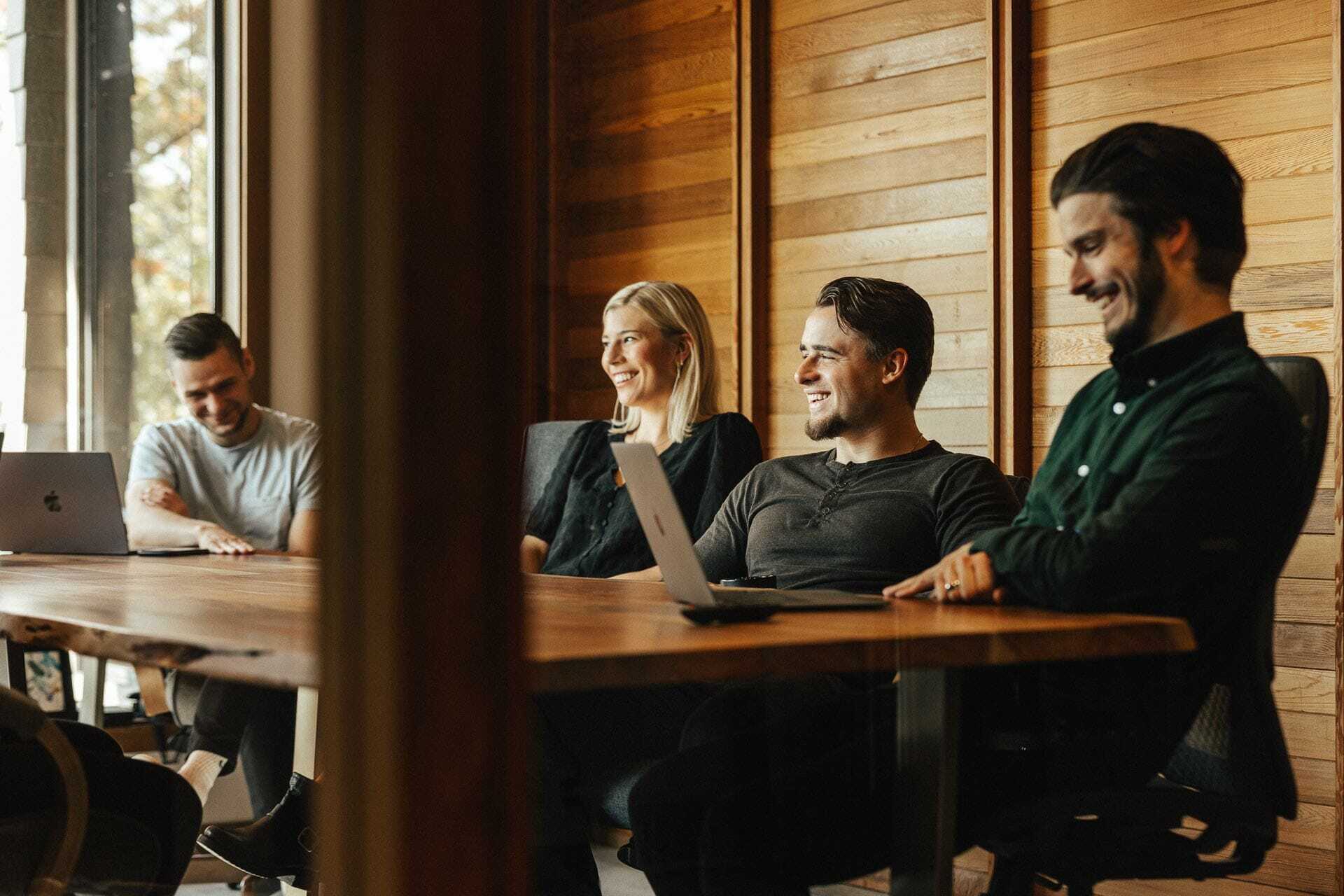 Four people seated around a wooden conference table, smiling and engaging in conversation. Two laptops are visible on the table. The setting appears to be a modern meeting room with wooden walls and large windows.