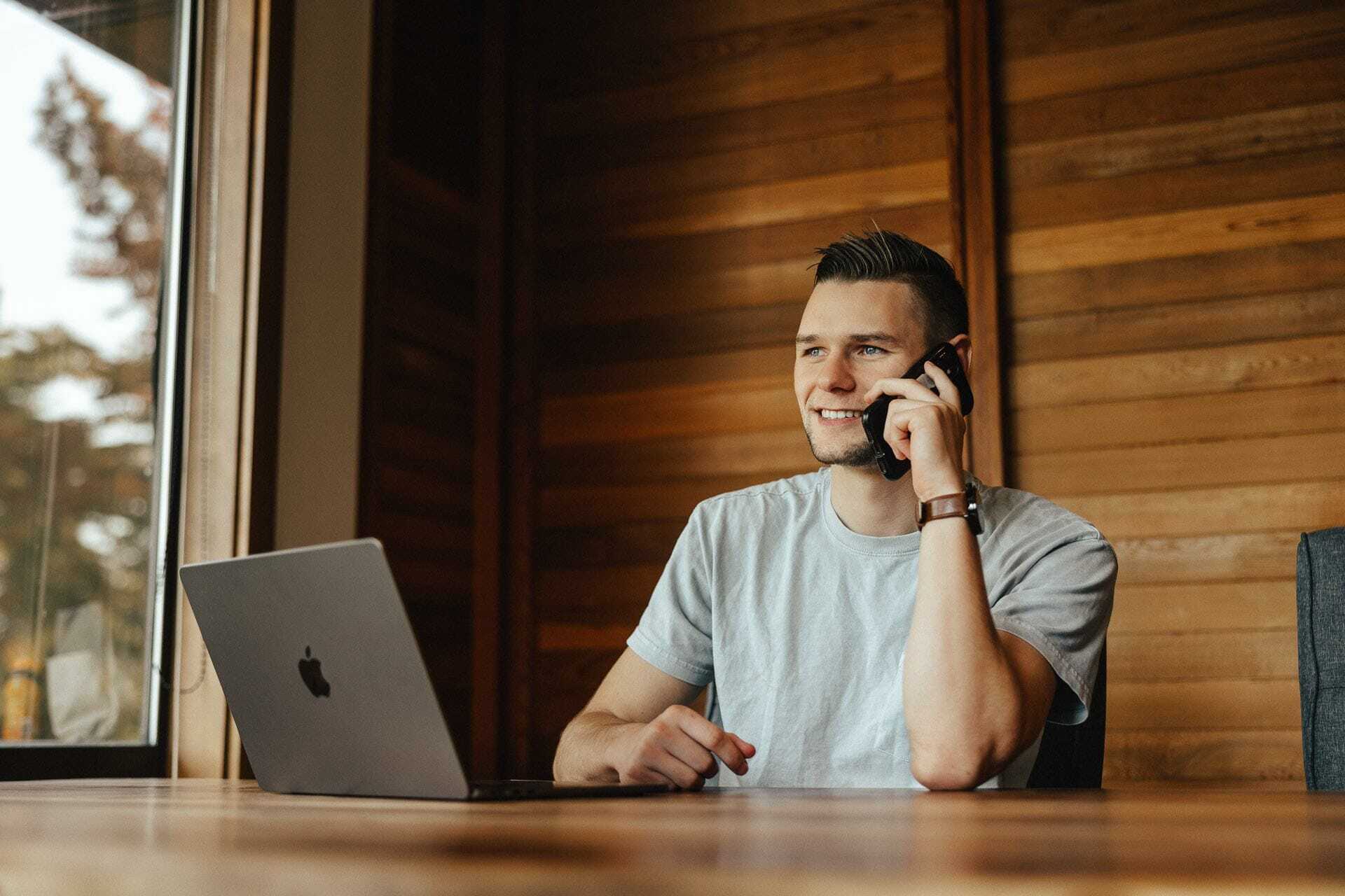 A person wearing a white T-shirt sits at a wooden table, talking on a smartphone. A laptop is open in front of them. The background features wooden walls and a large window showing trees outside.