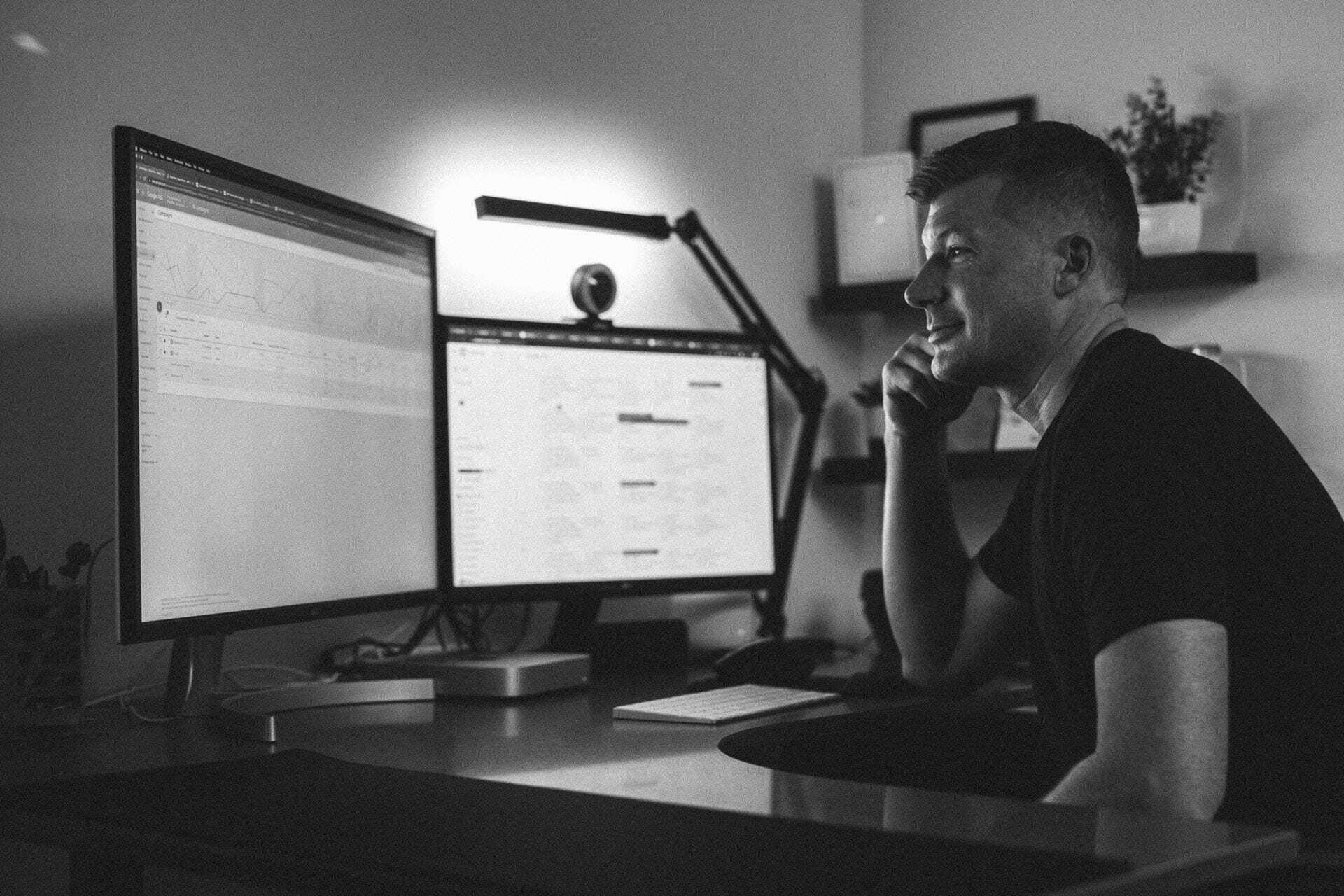 A person sitting at a desk, looking at two computer monitors displaying charts and data. The room is dimly lit with a desk lamp. Shelves with decor items are visible in the background. The scene is in black and white.