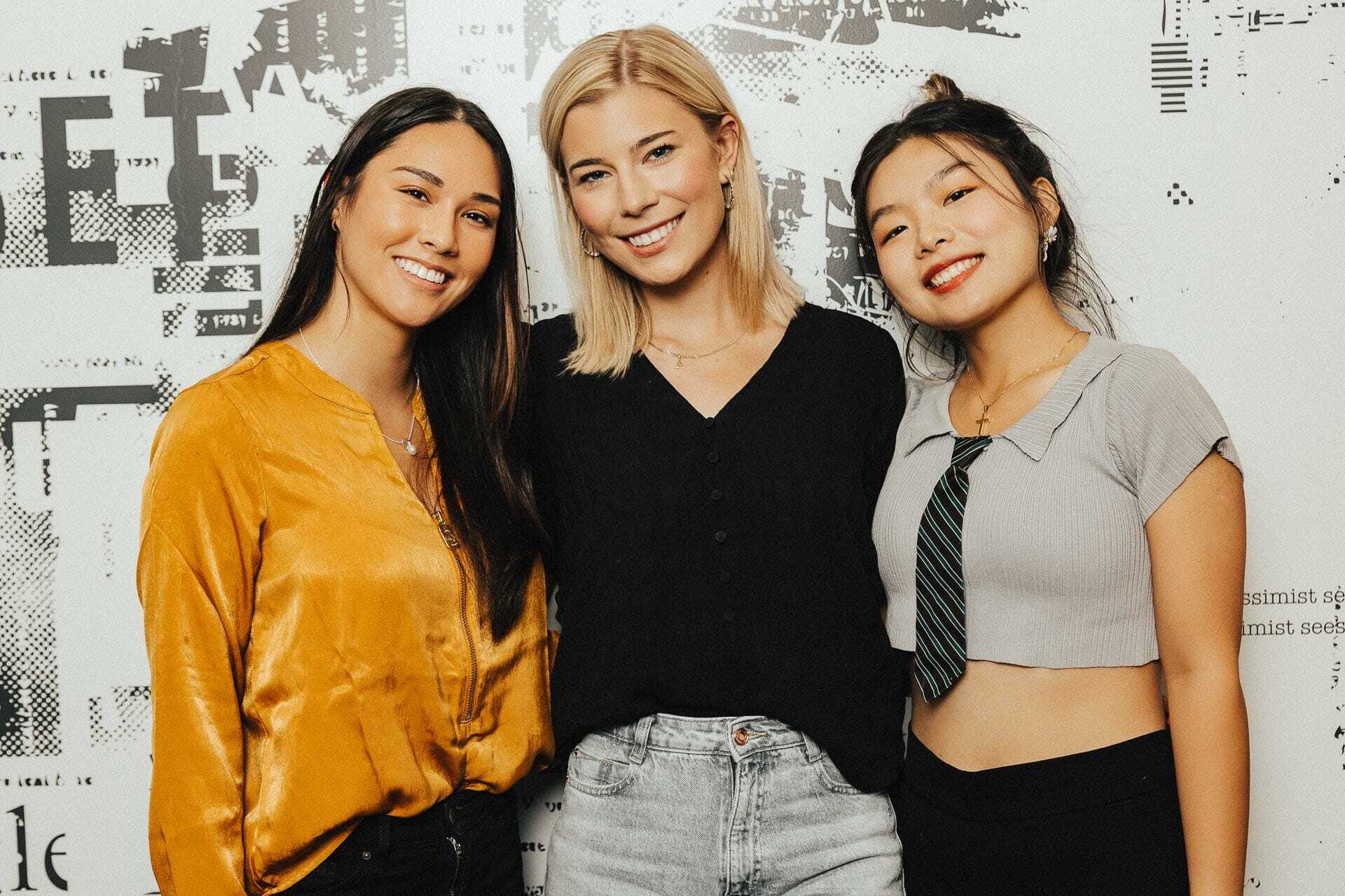 Three women are standing and smiling in front of a patterned wall. The woman on the left wears a yellow blouse, the middle woman wears a black blouse and light jeans, and the woman on the right wears a gray top with a tie.