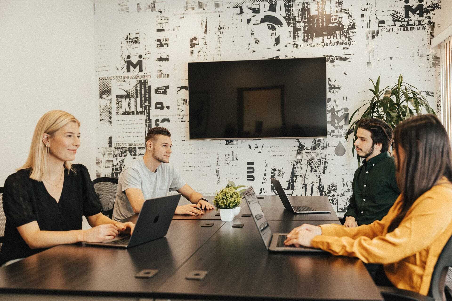 Four people are sitting around a conference table with laptops, engaged in a meeting. A large screen is mounted on the wall behind them, and a plant is in the corner. The room has a modern, casual vibe with graphic wallpaper.