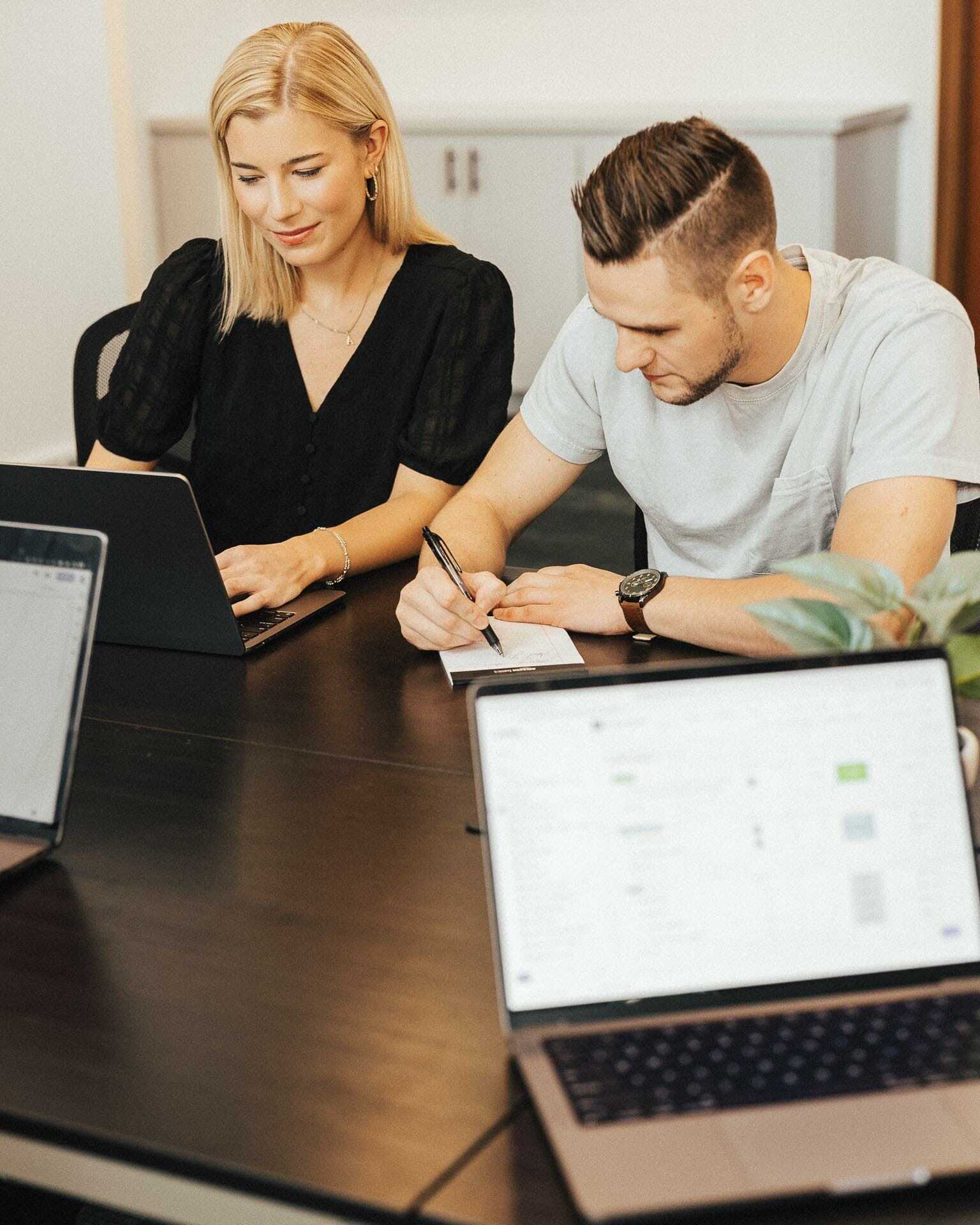 Two people working at a table with laptops. A woman is typing on her laptop, while a man takes notes on paper. Other laptops are visible on the table, along with a small plant in the foreground.