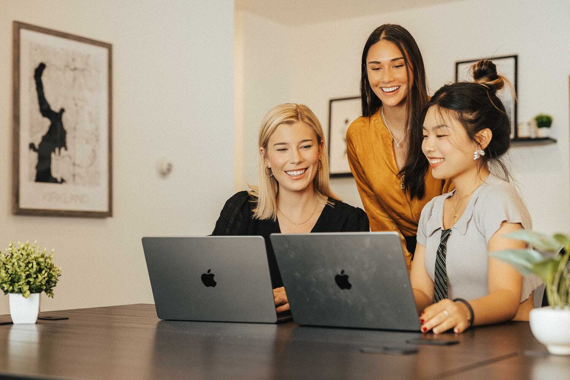Three women are at a table, smiling and looking at two laptops. One woman stands while the other two are seated. The room has minimal decor with artwork on the walls and small plants on the desk.