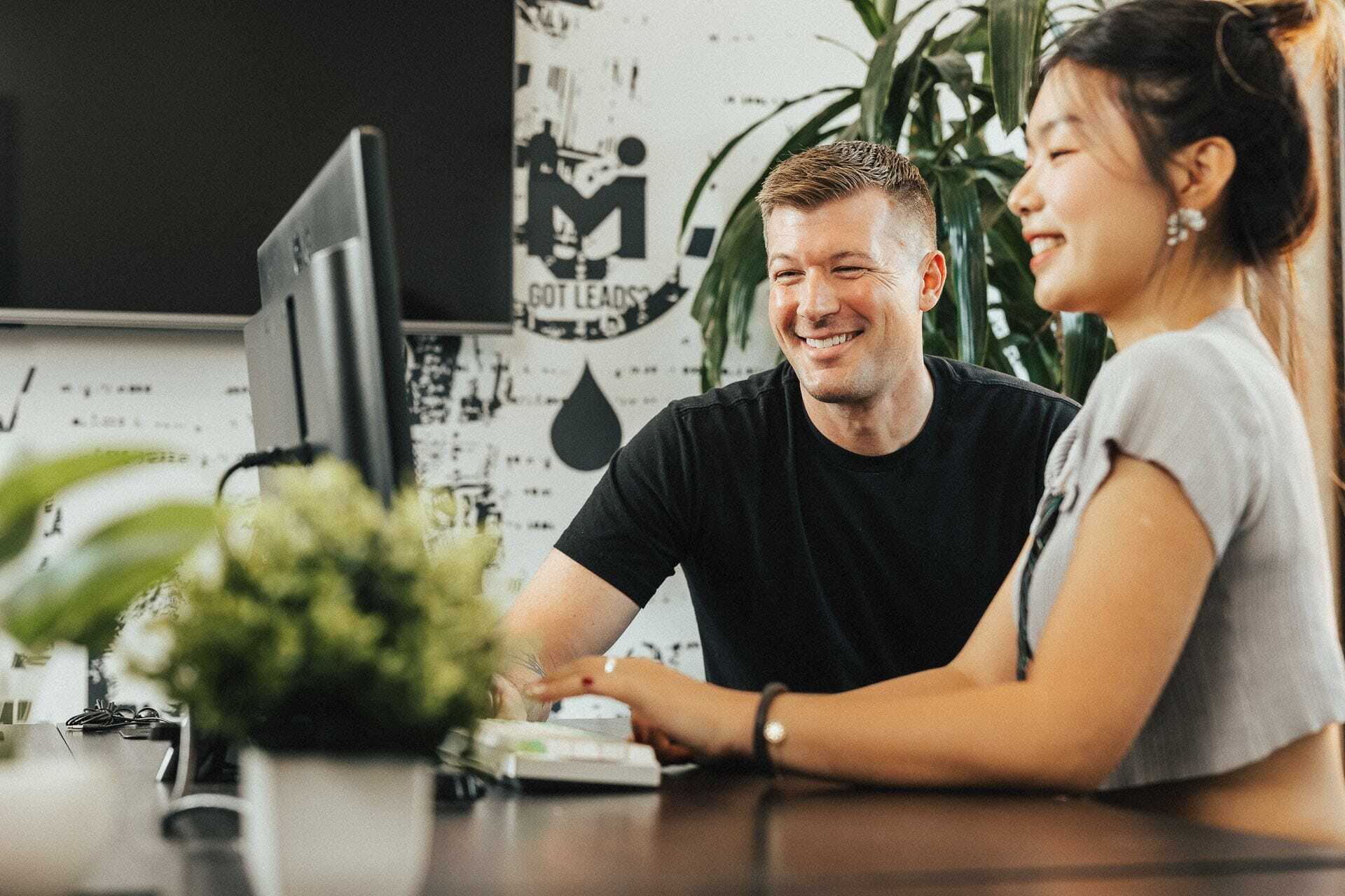 A man and a woman are smiling and looking at a computer screen together in an office setting. The desk is adorned with a small potted plant. The background features patterned wall decor and greenery.