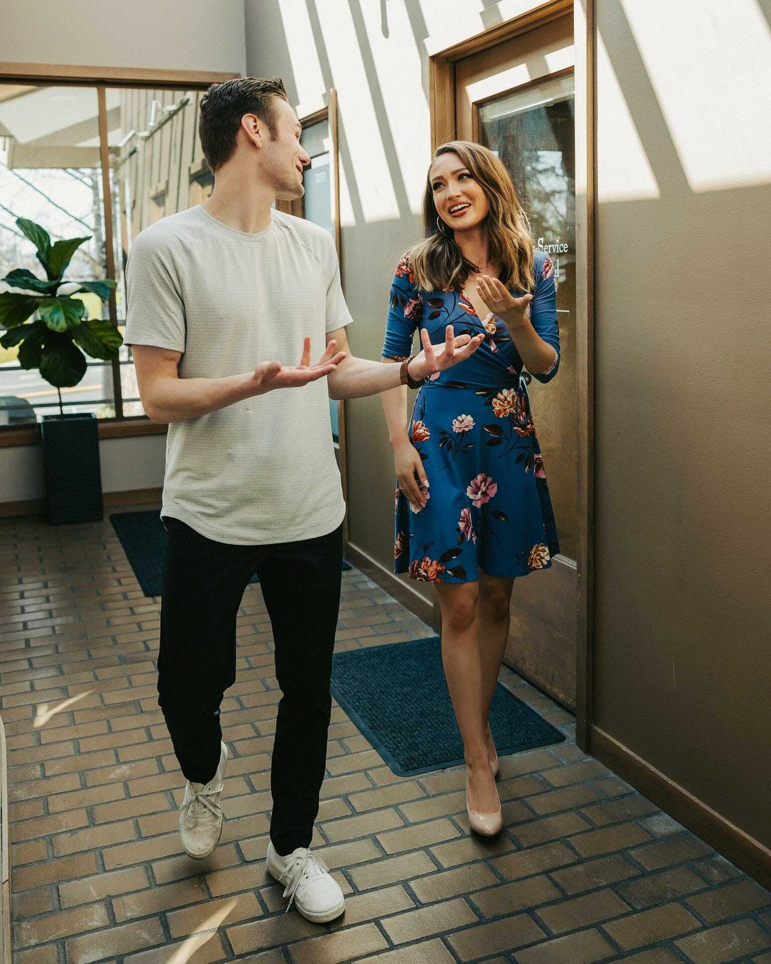 A man in a white t-shirt and black pants walks alongside a woman in a blue floral dress in a hallway with brick flooring. They are engaged in conversation. Sunlight streams in through the windows, illuminating a potted plant in the corner.