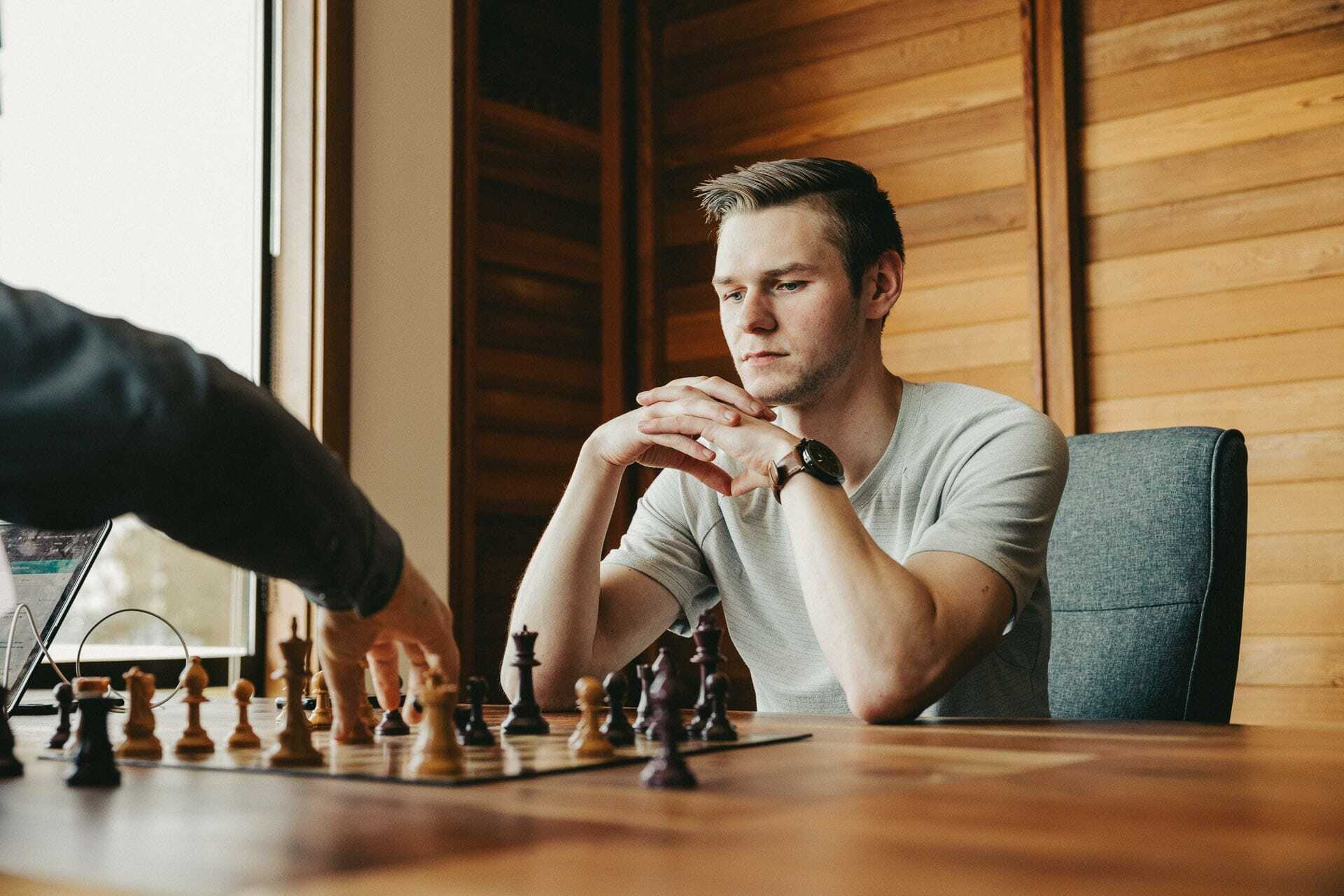 A person thoughtfully watches a chess game, hands clasped under their chin. They sit at a wooden table in a room with wooden panel walls. Sunlight filters through a window beside them.