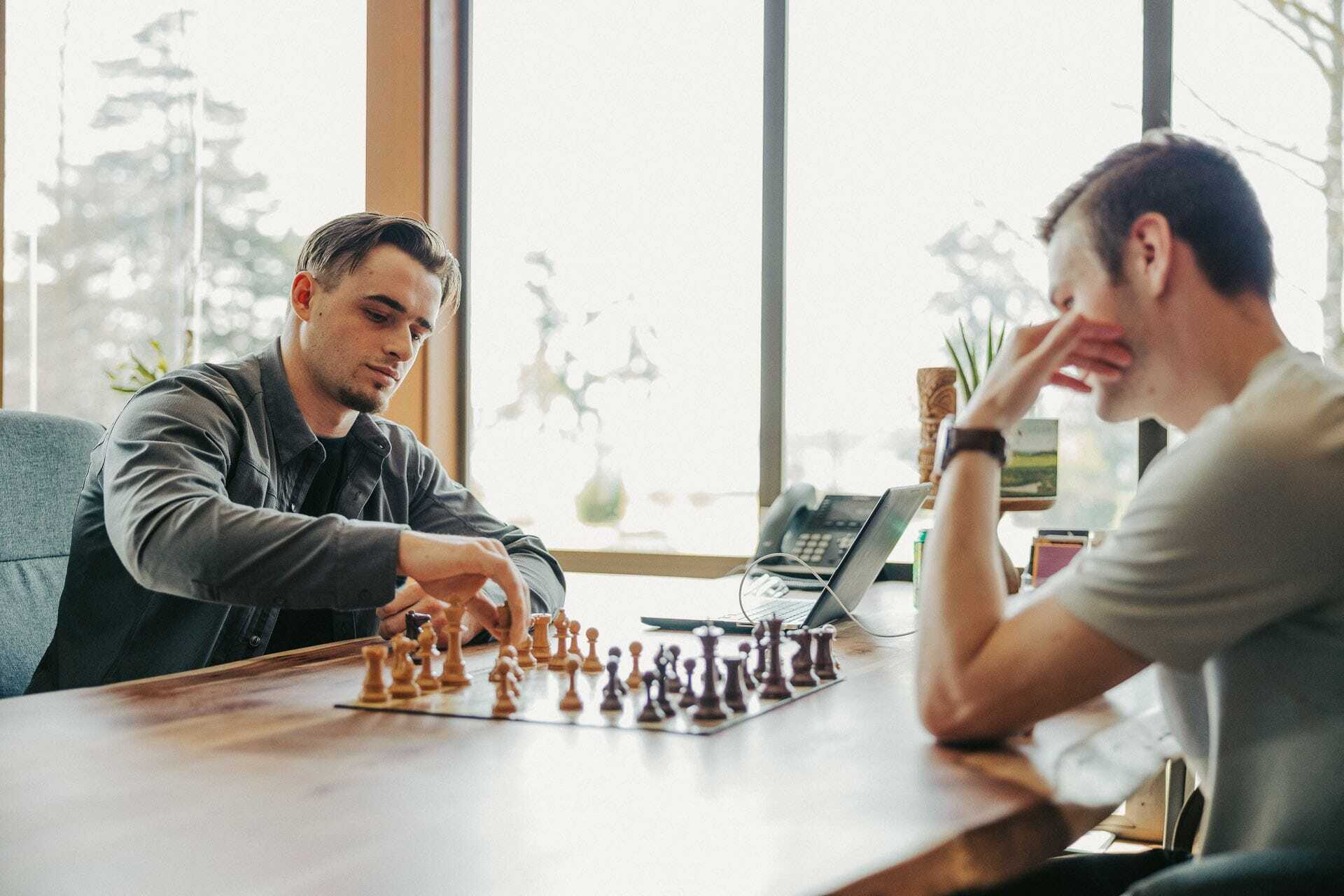 Two people sit at a wooden table playing chess. One person is making a move while the other watches intently. The room is well-lit with large windows in the background and some plants around.