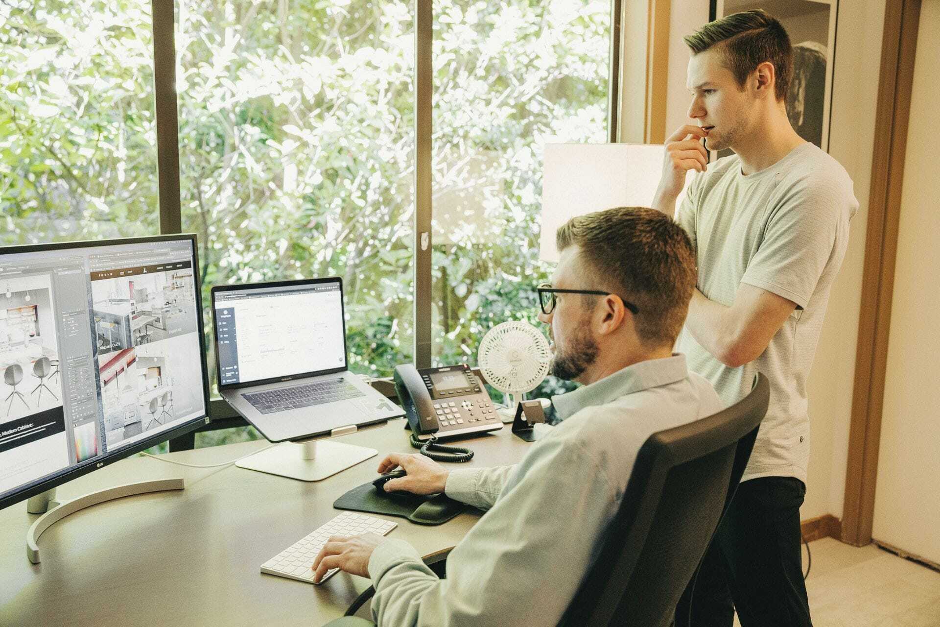 Two men work at a desk with multiple monitors and a laptop. One sits and uses a keyboard and mouse, while the other stands, looking at the screens. A window showing greenery is in the background.