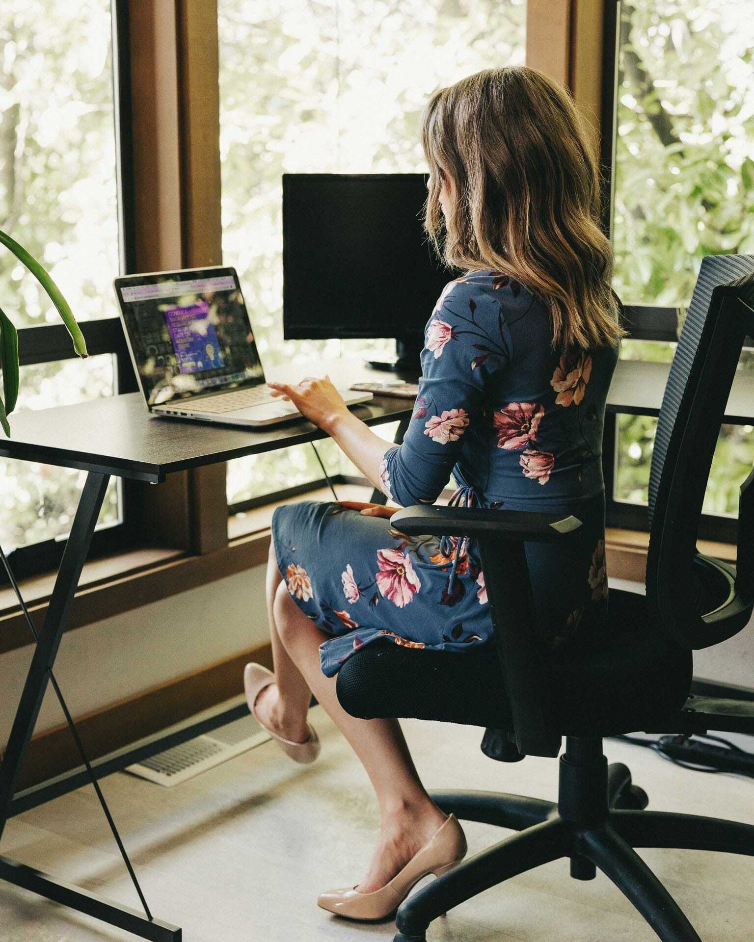 A woman in a floral dress sits in an office chair, working on a laptop at a desk. Two monitors are on the desk, and large windows show greenery outside. The setting is bright and well-lit.