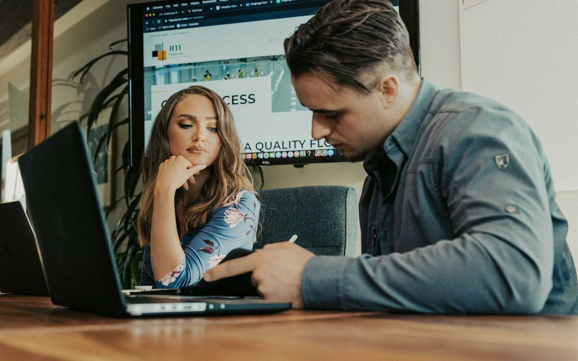 A woman and a man are sitting at a conference table with laptops. The woman is looking at the man, who is writing in a notebook. A large screen in the background displays a website. The setting appears to be an office.