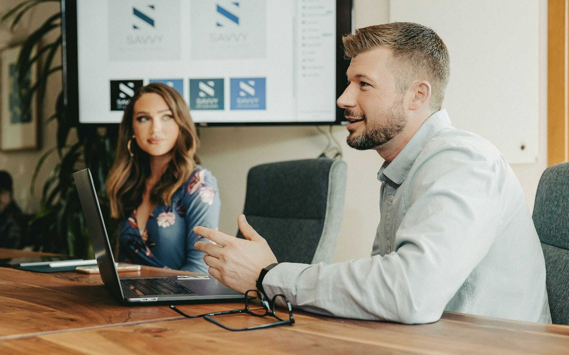 A man gives a presentation in an office setting, pointing at a laptop screen. A woman sits beside him, listening attentively. The background features a screen displaying the word SAVVY with a logo.
