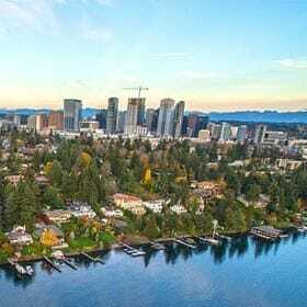Aerial view of a cityscape featuring a mix of tall skyscrapers and a waterfront residential area. Trees with autumn foliage surround the houses, and a calm body of water reflects the clear blue sky. Distant mountains complete the horizon.